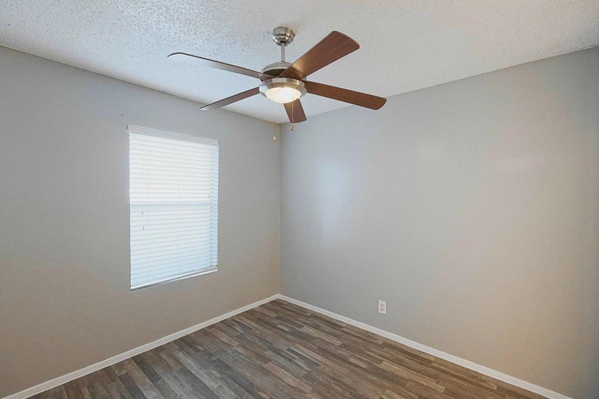 A plain, empty room featuring a light-colored wall, a ceiling fan with wooden blades, and a window with blinds. The flooring is made of wooden planks, creating a simple and spacious atmosphere suitable for various uses.