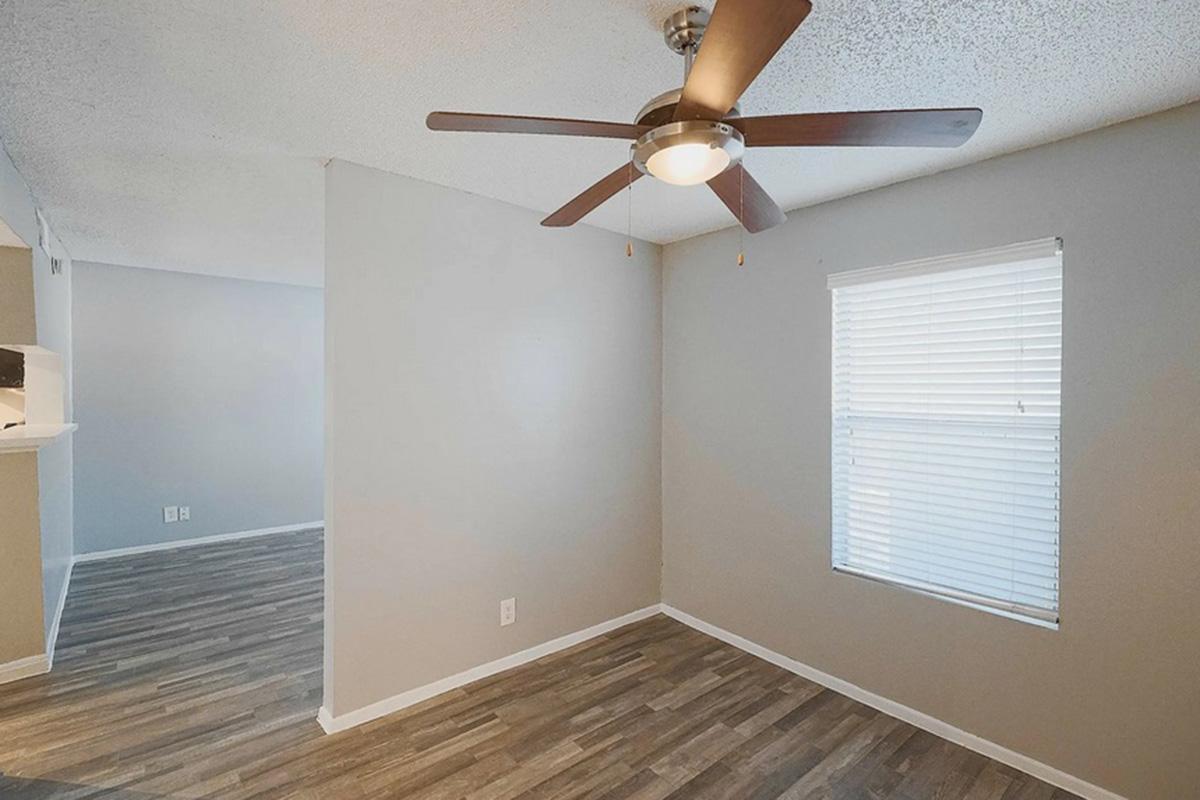 A spacious room featuring light gray walls, a ceiling fan with wooden blades, and a window with white blinds. The floor is covered with light-colored laminate. The layout is open, leading to adjoining areas.