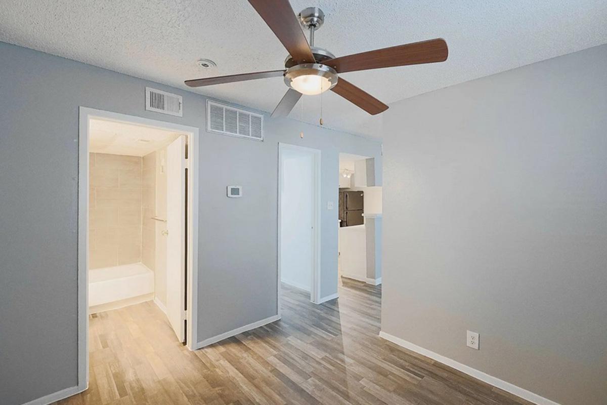 A modern interior living space featuring a ceiling fan with wooden blades, light gray walls, and laminate flooring. A doorway on the left leads to a bathroom, while an open area beyond it connects to a kitchen. Bright lighting enhances the clean, contemporary atmosphere.