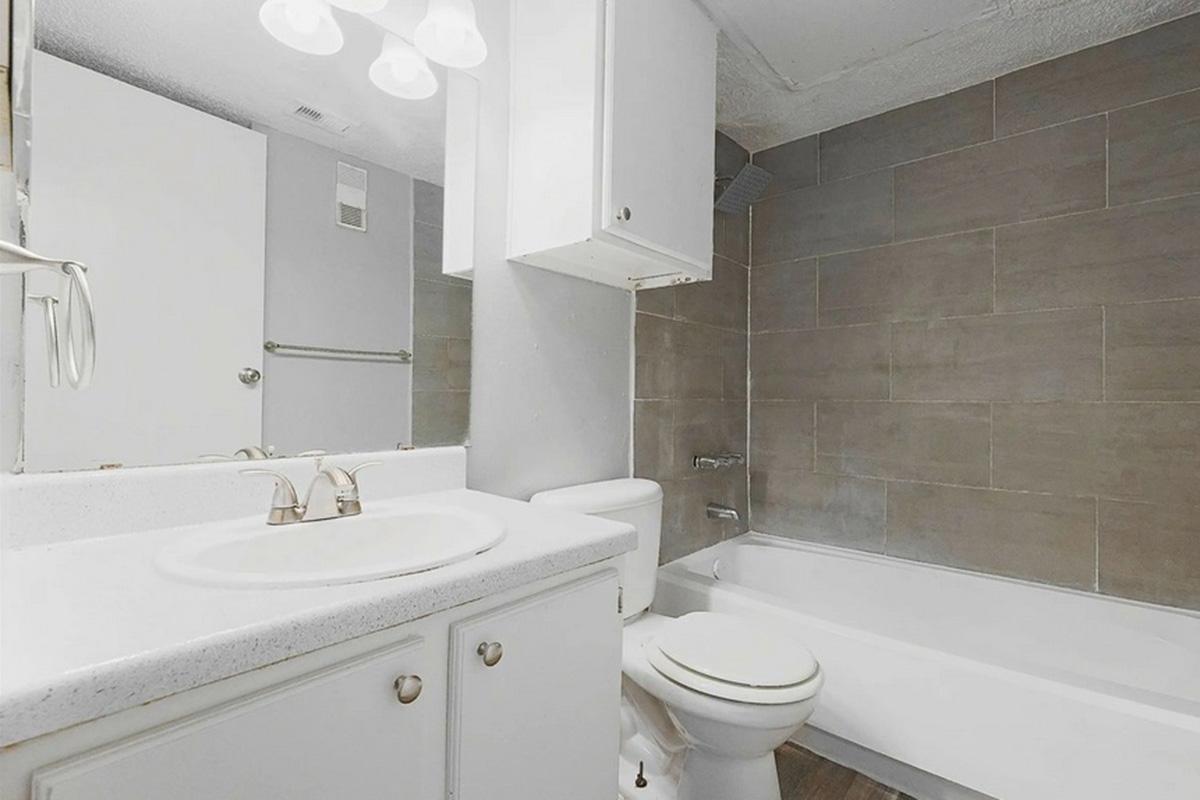A clean and modern bathroom featuring a white sink with two faucets, a toilet, and a bathtub. The walls are tiled in gray, and there is a well-lit mirror above the sink. The flooring is a light wood, creating a bright and spacious atmosphere.