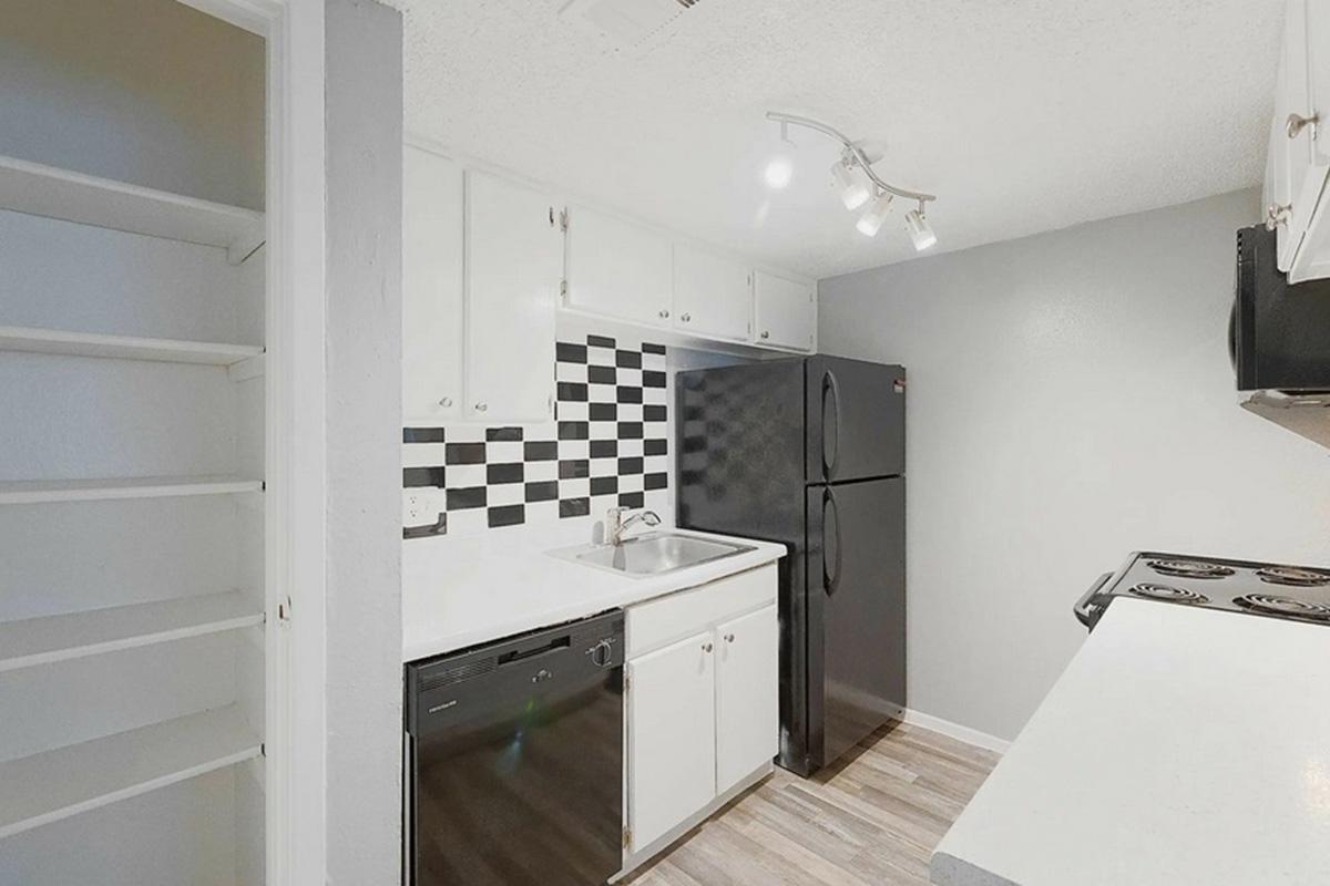 A modern kitchen featuring white cabinetry, a black refrigerator, a black and white checkered backsplash, and a stove. The space includes a sink and dishwasher, with overhead track lighting illuminating the area. Light wood flooring adds warmth to the contemporary design.