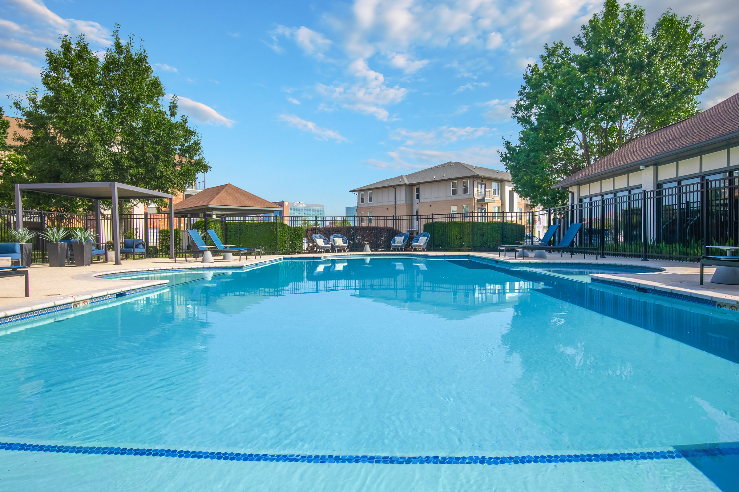 A clear swimming pool surrounded by lounge chairs and shaded seating areas. Green trees and a bright blue sky create a serene atmosphere. Adjacent buildings are in the background, adding to the residential ambiance of the setting.