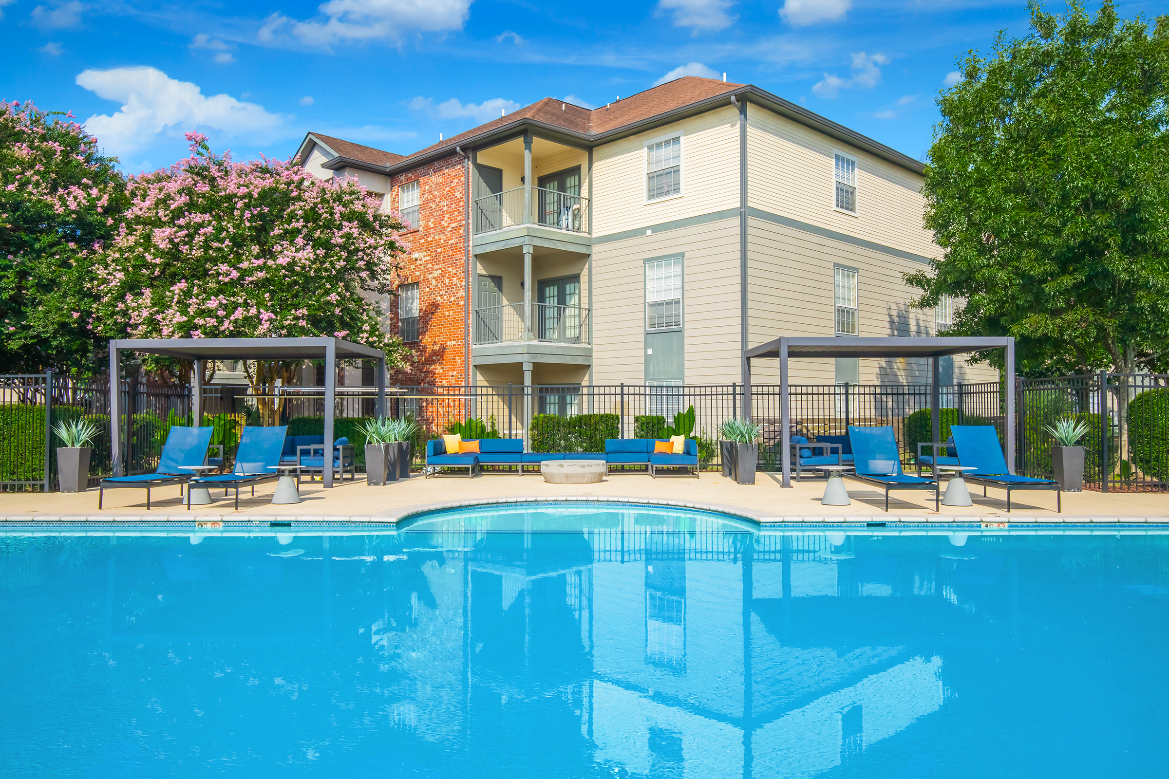 A clear blue swimming pool with chaise lounges and shaded cabanas. Surrounding the pool is greenery and flowering trees. In the background, a three-story apartment building features a mix of brick and siding. The sky is blue with scattered clouds, creating a bright and inviting atmosphere.