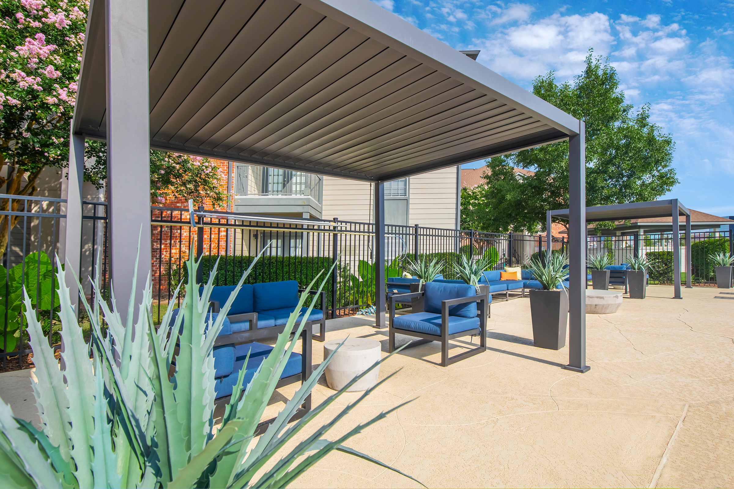 Outdoor lounge area featuring modern seating under shaded pergolas. The space is adorned with potted plants and surrounded by green landscaping, set against a clear blue sky.