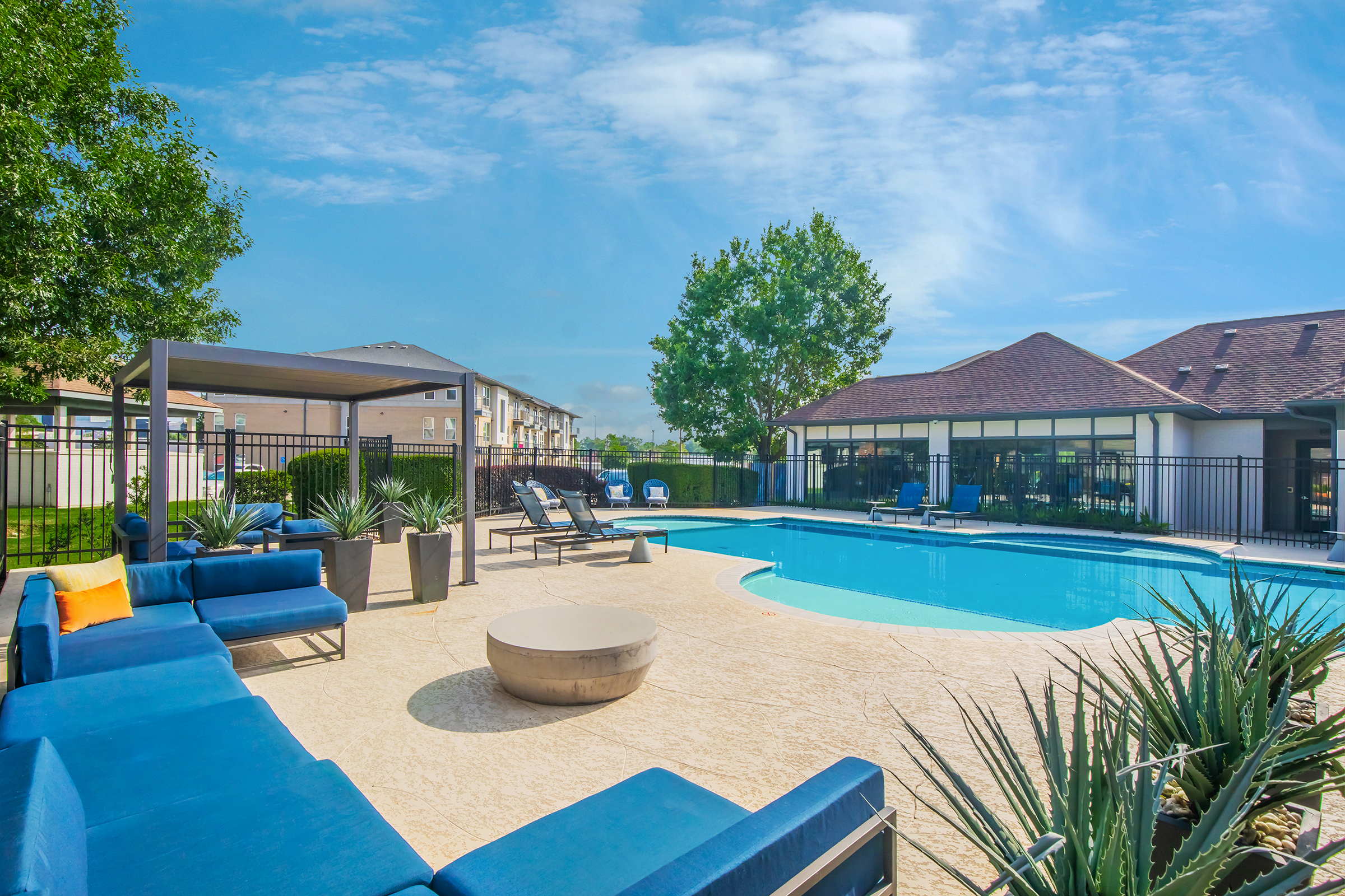 A spacious outdoor pool area featuring a blue lounge seating arrangement, a round fire pit, and lush greenery. The pool is surrounded by a safety fence and includes a shaded pergola, with nearby lounges and palm-like plants. Clear blue sky with scattered clouds enhances the inviting atmosphere.