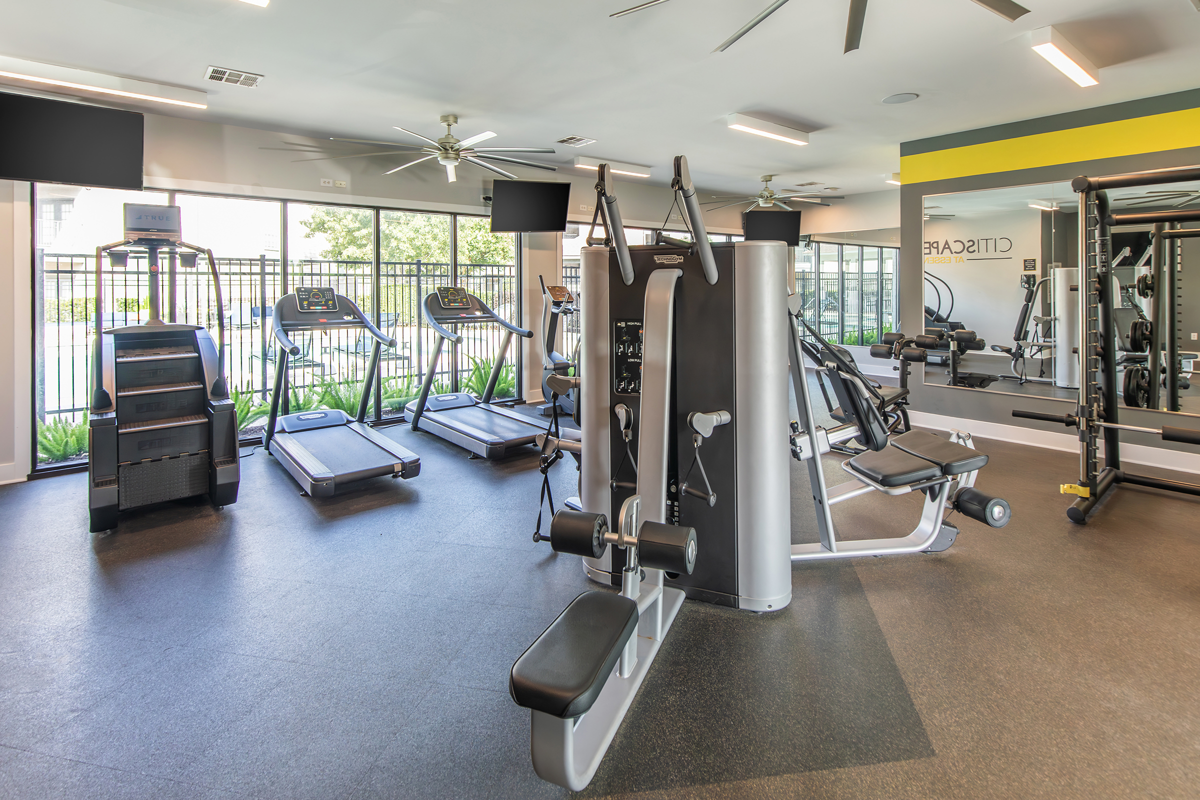 A modern gym interior featuring various fitness equipment including treadmills, a multi-station weight machine, and large windows providing natural light. The floor is covered with rubber tiles, and there are ceiling fans and wall-mounted TVs for entertainment. Greenery is visible outside the windows.