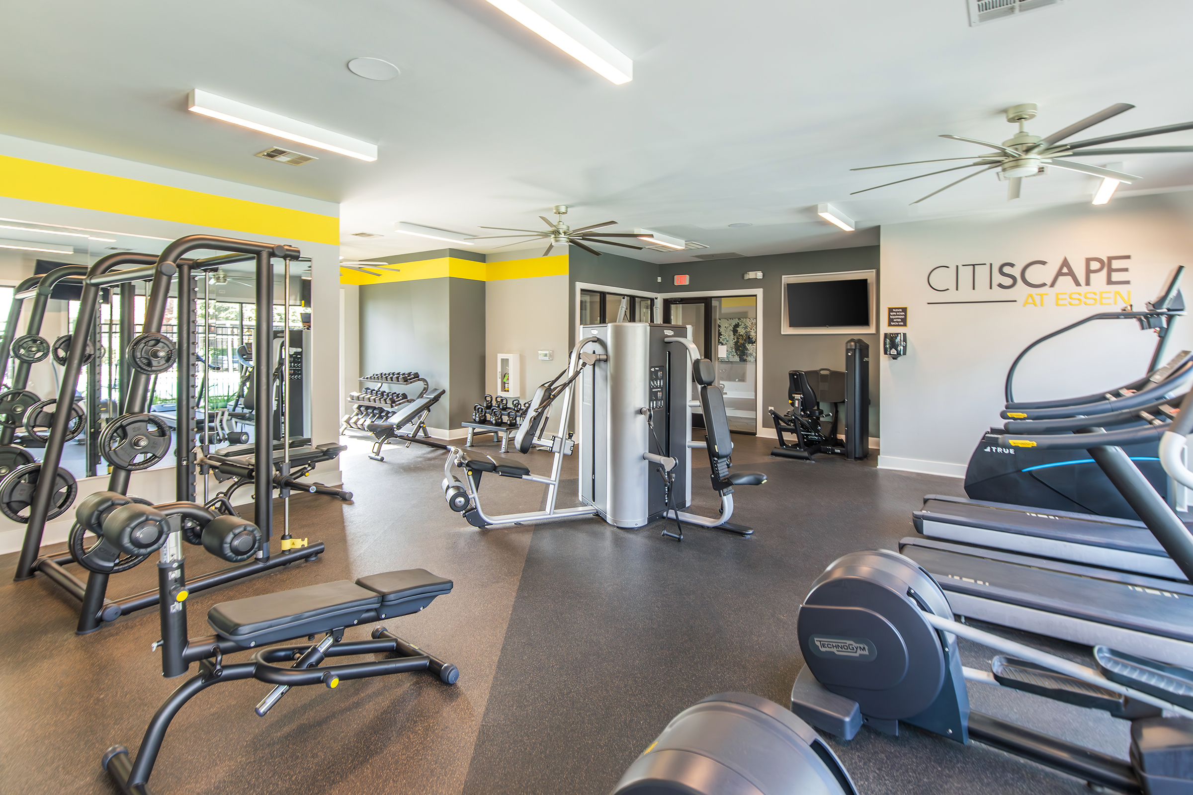 A modern gym interior featuring various exercise equipment, including weight machines, free weights, and cardio machines. The space has bright yellow accents on the walls, large mirrors, and ceiling fans, creating a welcoming environment for fitness activities.