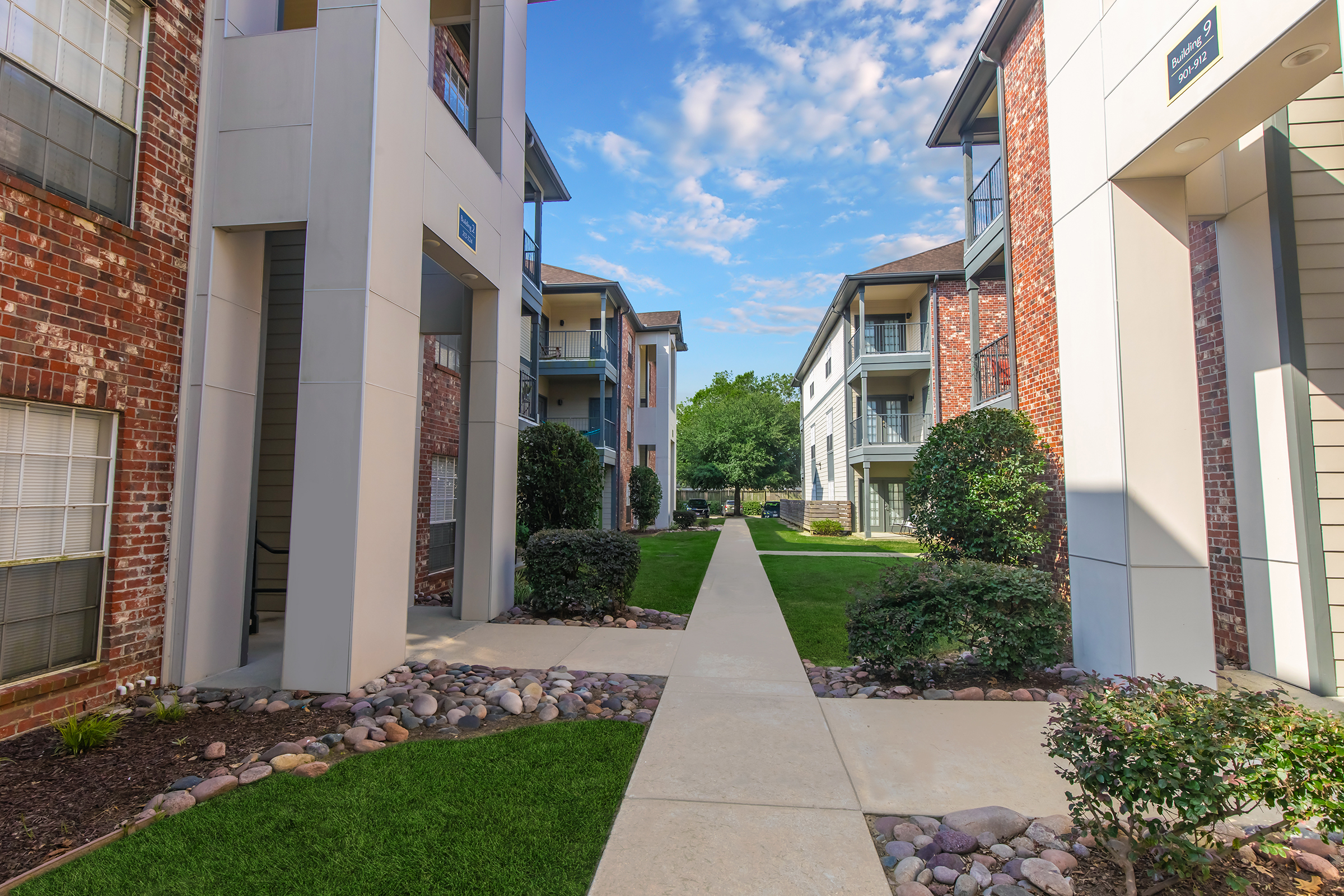 A well-maintained apartment complex featuring two buildings on either side of a paved walkway. Lush green grass, shrubs, and small trees line the path, with blue skies and scattered clouds overhead, creating a serene and inviting atmosphere.