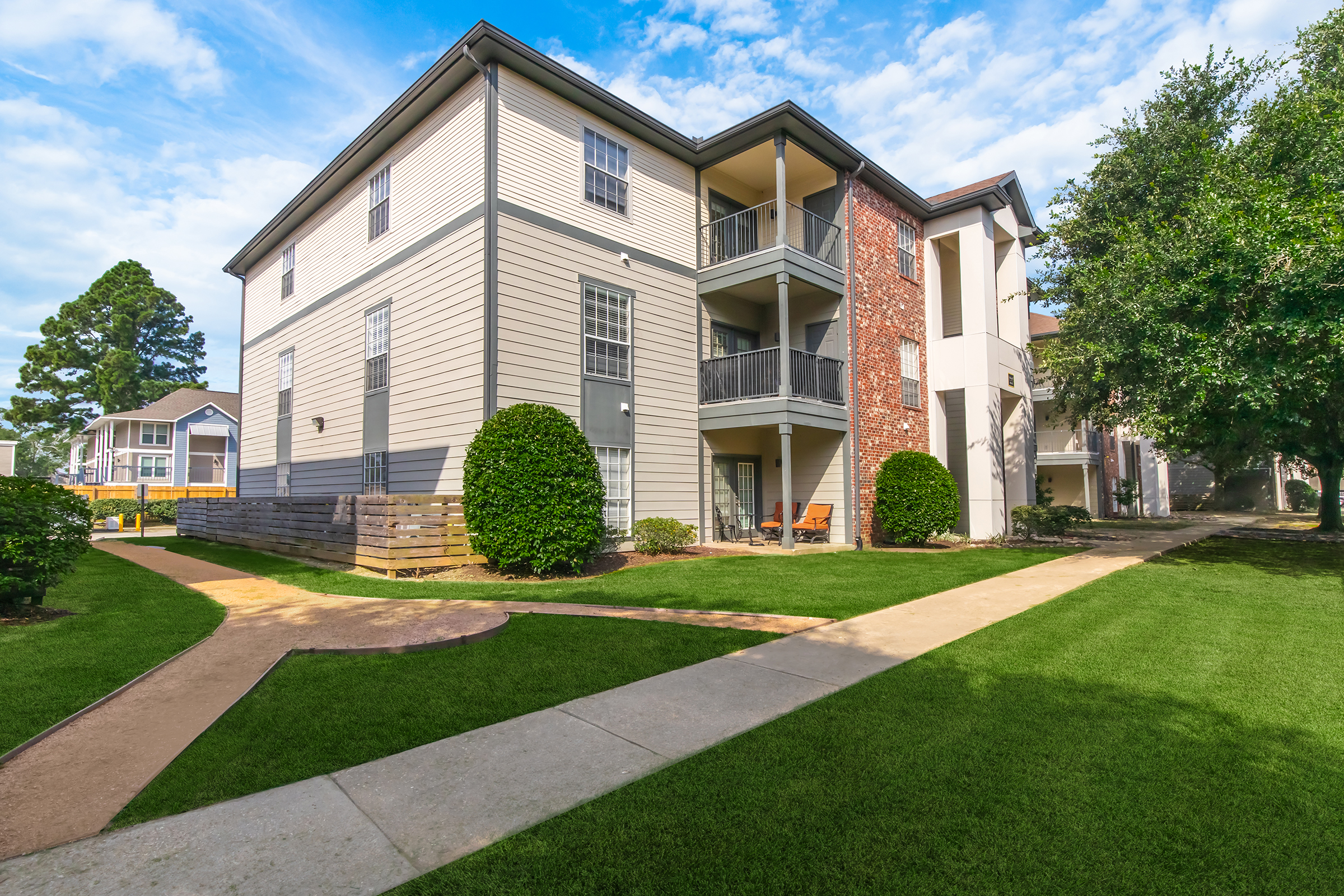 A well-maintained apartment complex with a mix of brick and siding exteriors. The building features multiple balconies, surrounded by lush green lawns and landscaped pathways. Trees are visible in the background, creating a pleasant outdoor environment. Bright, sunny sky adds to the inviting atmosphere.