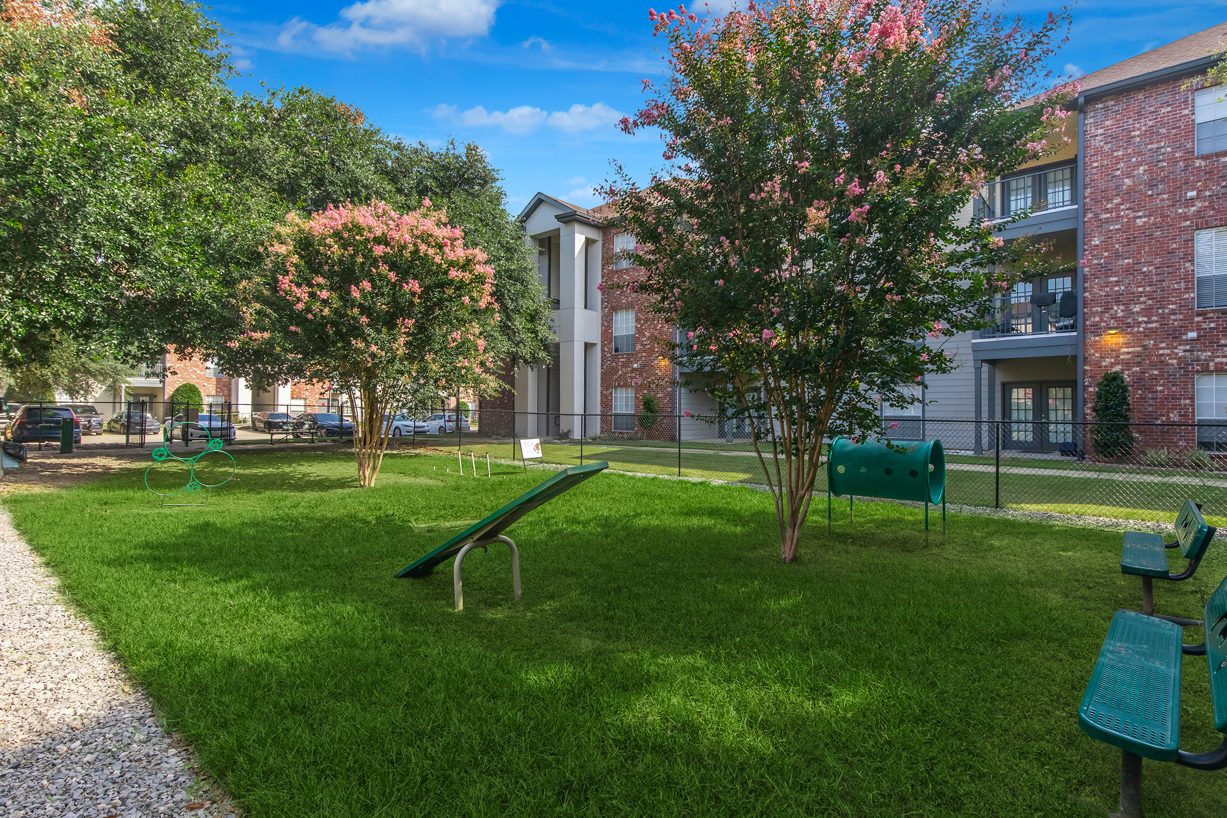 A well-maintained outdoor area with green grass, two flowering trees, and a playground structure. There are benches along the sides, a fence enclosing the space, and apartment buildings in the background under a clear blue sky.