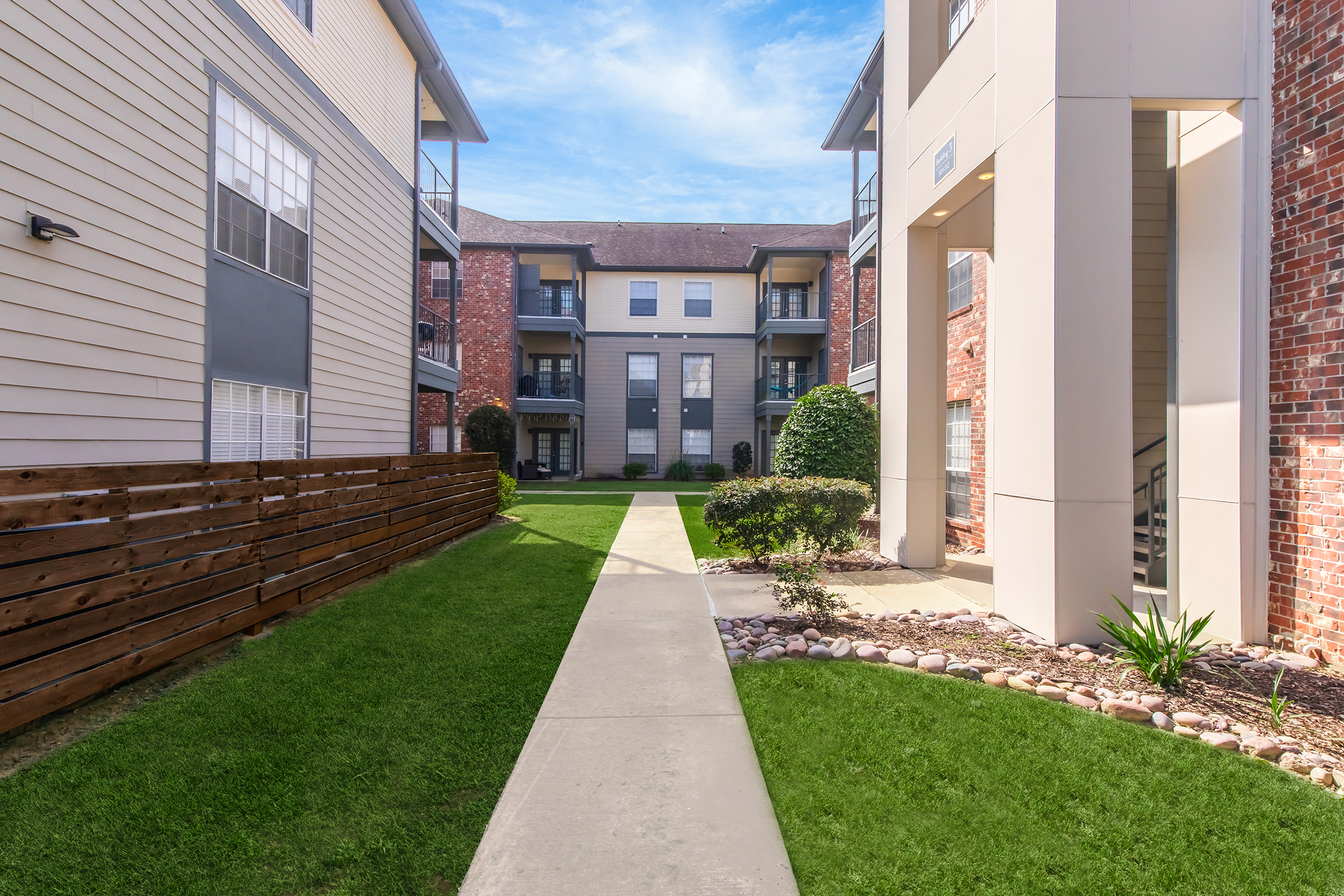 A view of a well-maintained apartment complex featuring a paved walkway surrounded by lush green grass, small bushes, and decorative stones. Two buildings with balconies and various window designs are visible, set against a blue sky with scattered clouds.