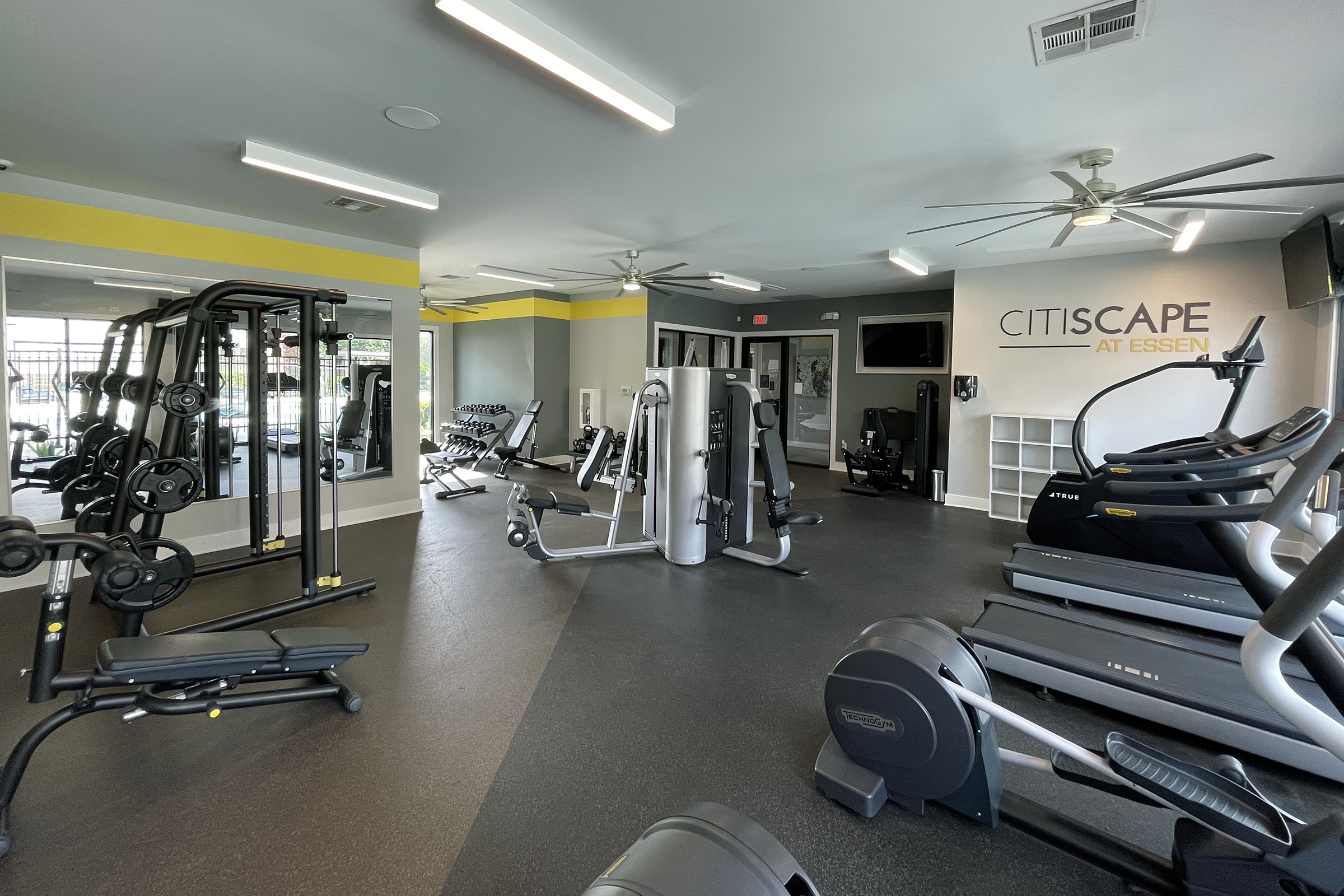 Interior view of a modern gym featuring various exercise equipment, including weight machines, cardio machines, and free weights. The space is well-lit with ceiling fans and large mirrors, and the wall displays the "CitiScape at Essen" logo. The flooring is dark and the walls are painted in light colors.