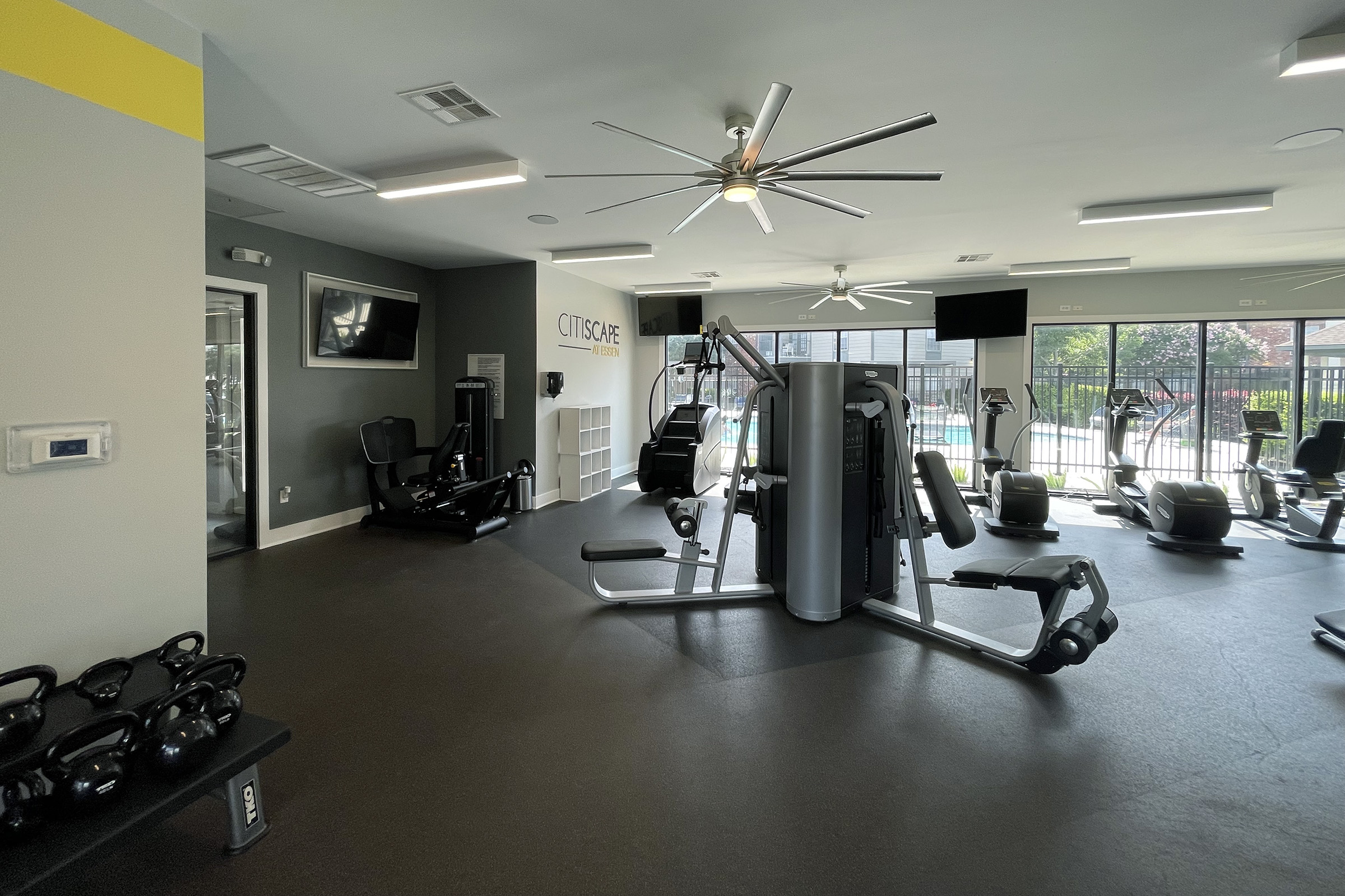 A spacious gym interior featuring various exercise equipment, including weight machines and cardio machines. Large windows offer natural light, and there are ceiling fans and flat-screen monitors. Dumbbells are organized on a rack in one corner.
