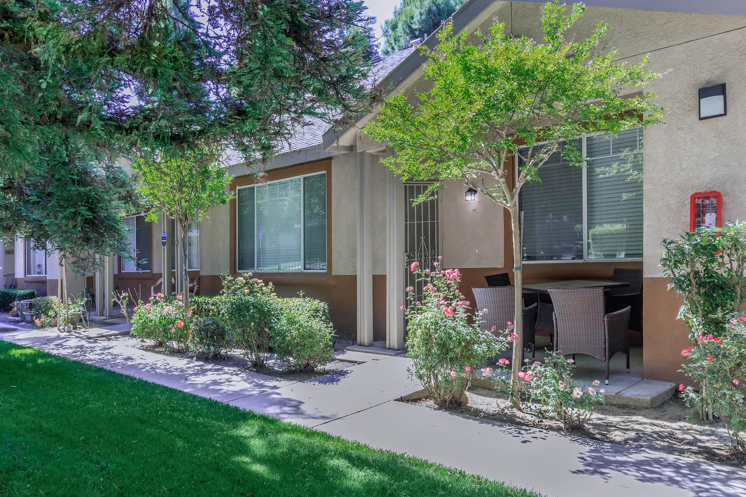 A well-maintained apartment building with a landscaped front yard, featuring green grass, shrubs, and rose bushes. The building has large windows, outdoor seating with wicker chairs, and a fire extinguisher visible on the wall. Sunlight casts gentle shadows on the path.