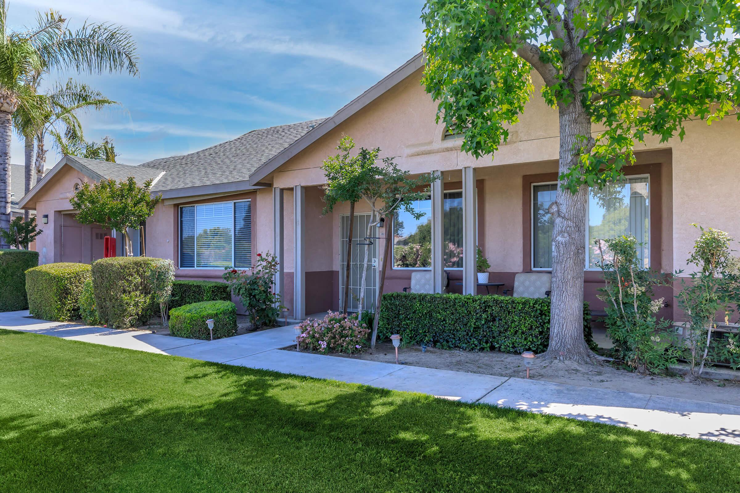 A well-maintained residential home with a manicured front yard featuring green grass and neatly trimmed bushes. The house has a light pink exterior, large windows, and a few potted plants. There are trees providing shade, and the setting has a clear blue sky.