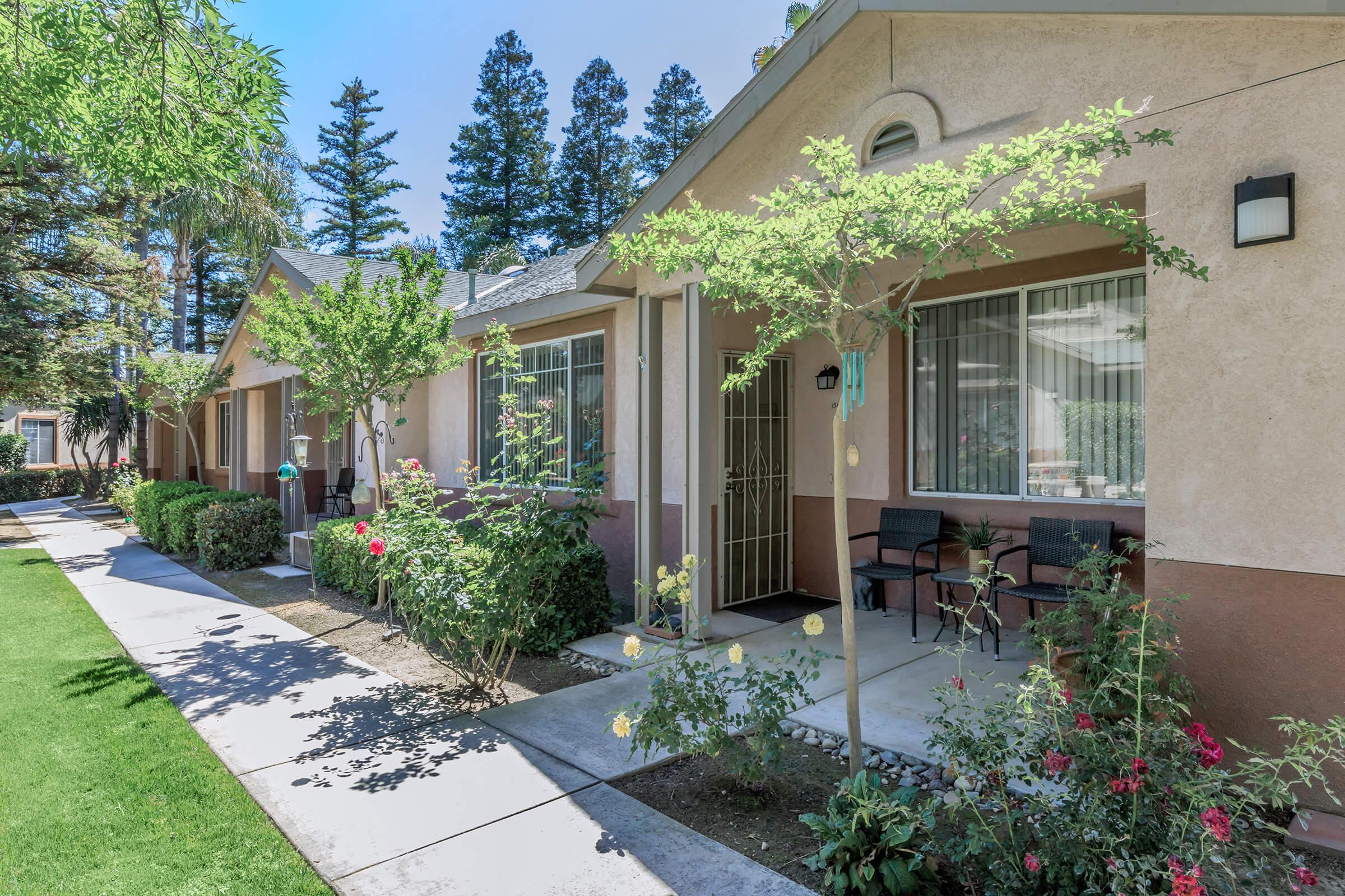 A row of residential buildings with a well-maintained lawn and flower beds. Each unit features large windows and a small porch area with chairs. Lush greenery and tall trees are visible in the background under a clear blue sky.