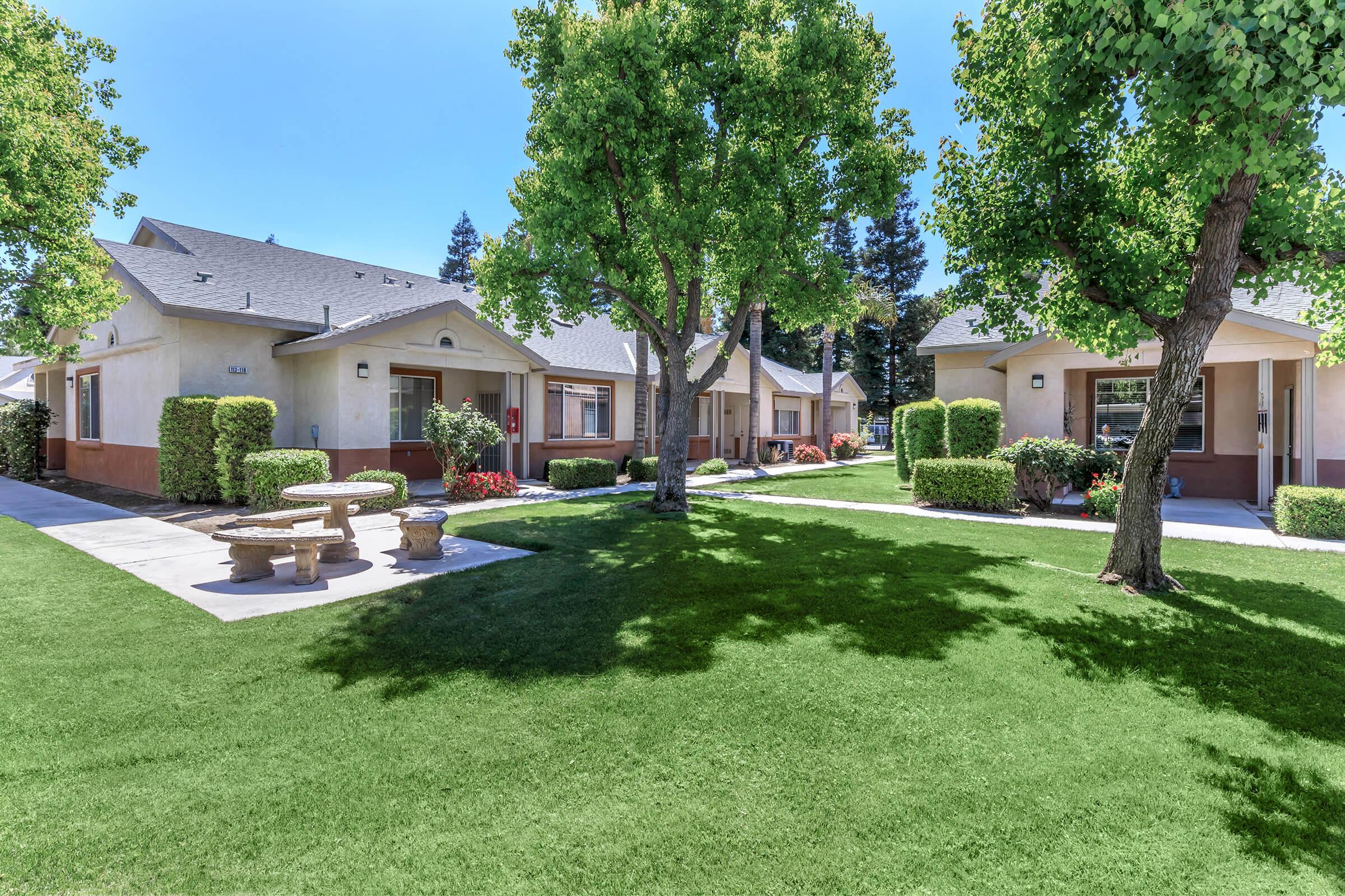 A well-maintained residential area featuring several single-story buildings surrounded by lush green lawns and trees. There is a stone seating area in the foreground and vibrant flower beds, creating a welcoming and pleasant environment. The sky is clear and bright, enhancing the appeal of the setting.