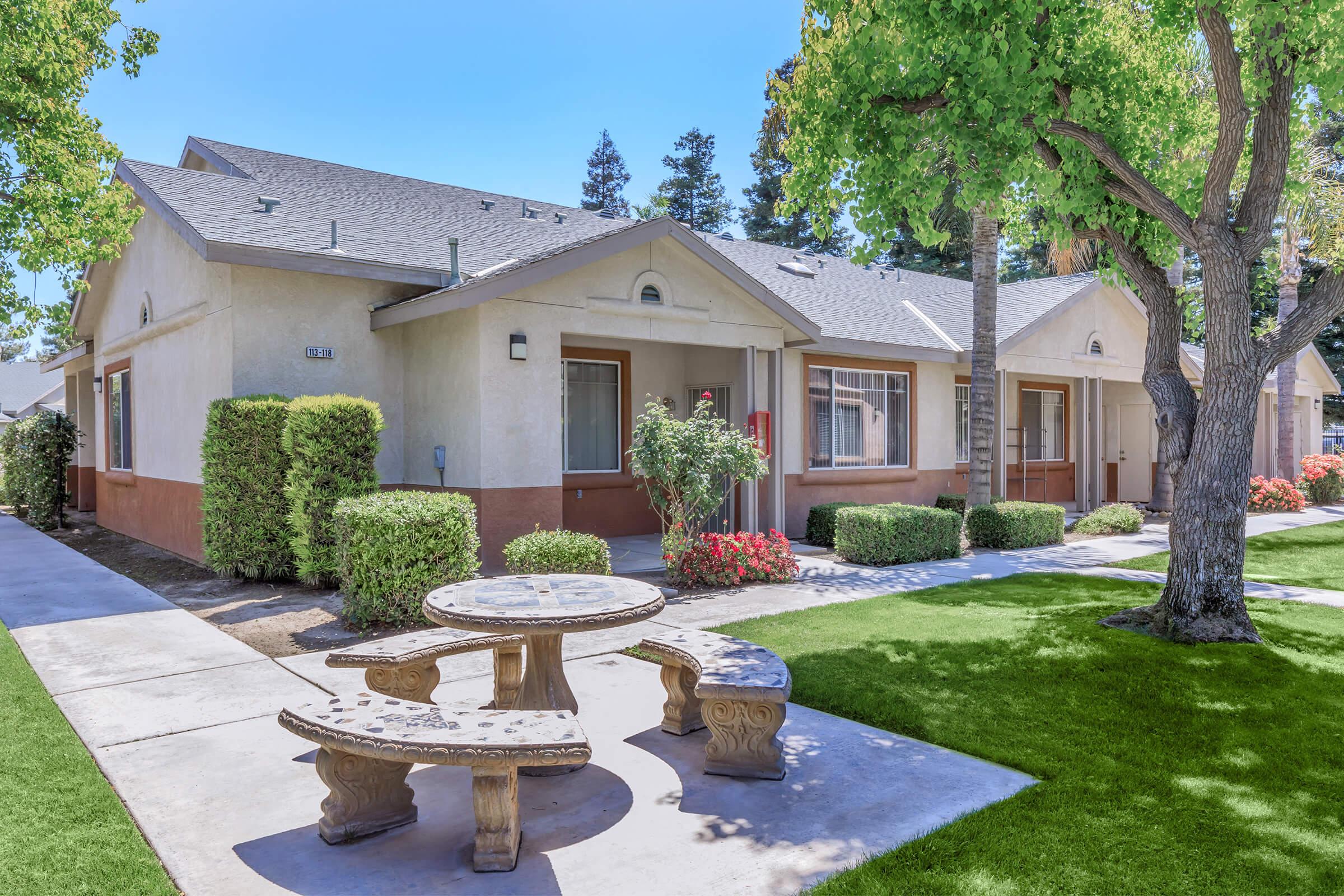 A row of beige single-story residential units with sloped roofs, surrounded by well-maintained landscaping including shrubs and trees. In the foreground, a stone picnic table sits on green grass, creating a welcoming outdoor space. Bright blue sky overhead, emphasizing a pleasant day.