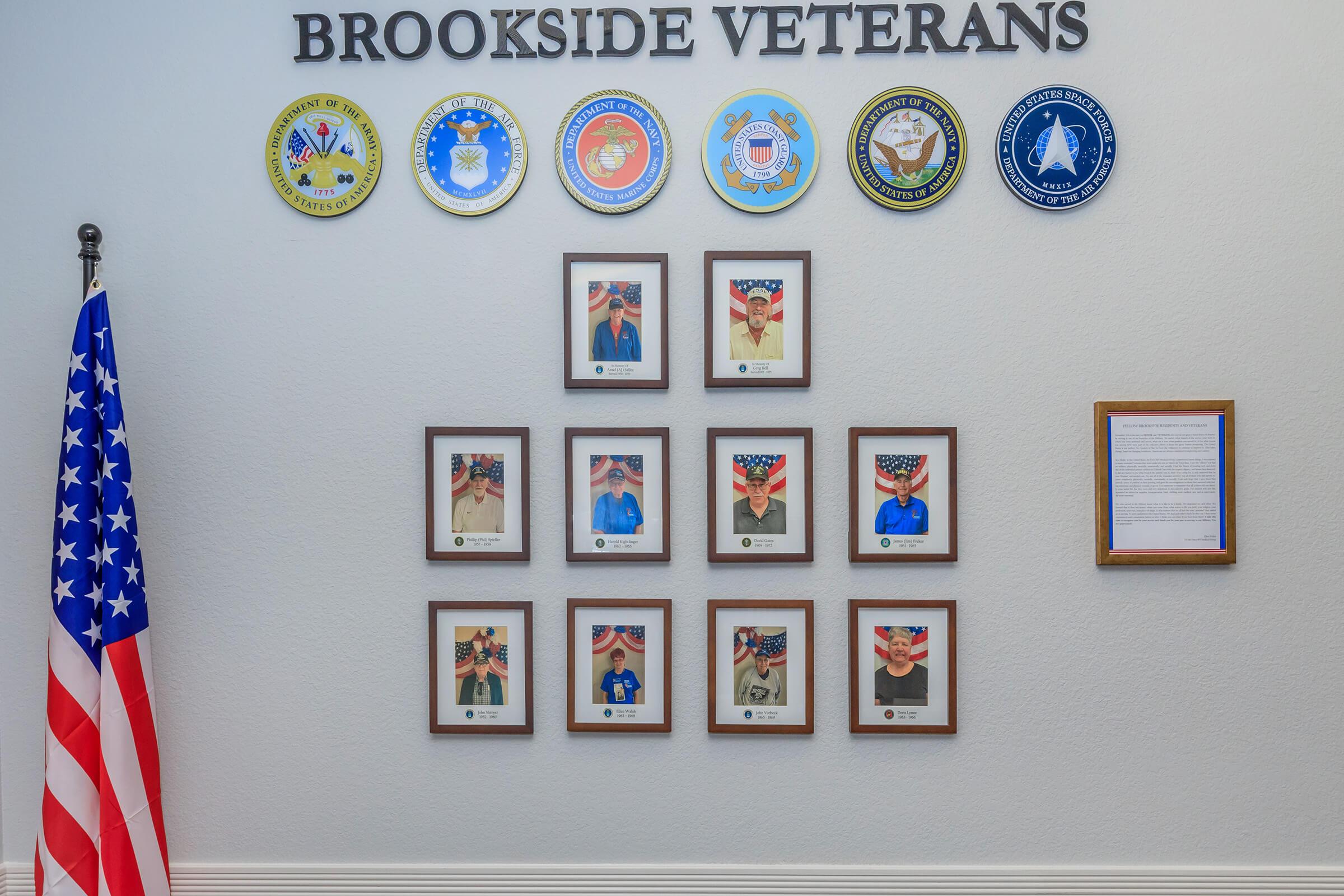 A wall display titled "Brookside Veterans" featuring various military service emblems at the top. Below, there are framed portraits of veterans, each with an American flag backdrop, showcasing their service. A U.S. flag stands to the left side of the display.