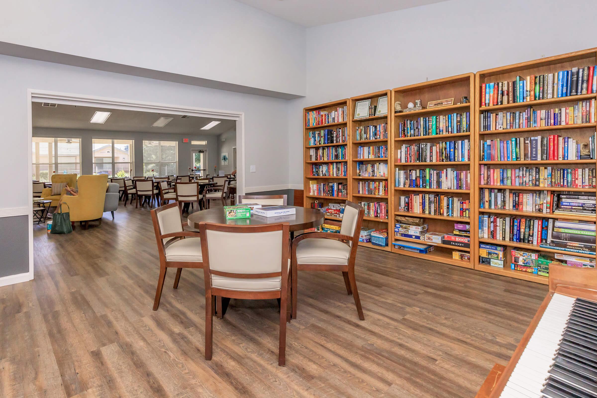 A spacious common area featuring a table with four chairs in the foreground, a bookshelf filled with various books along the wall, and a cozy seating area visible in the background with large windows allowing natural light. The flooring is wooden, creating a warm ambiance.
