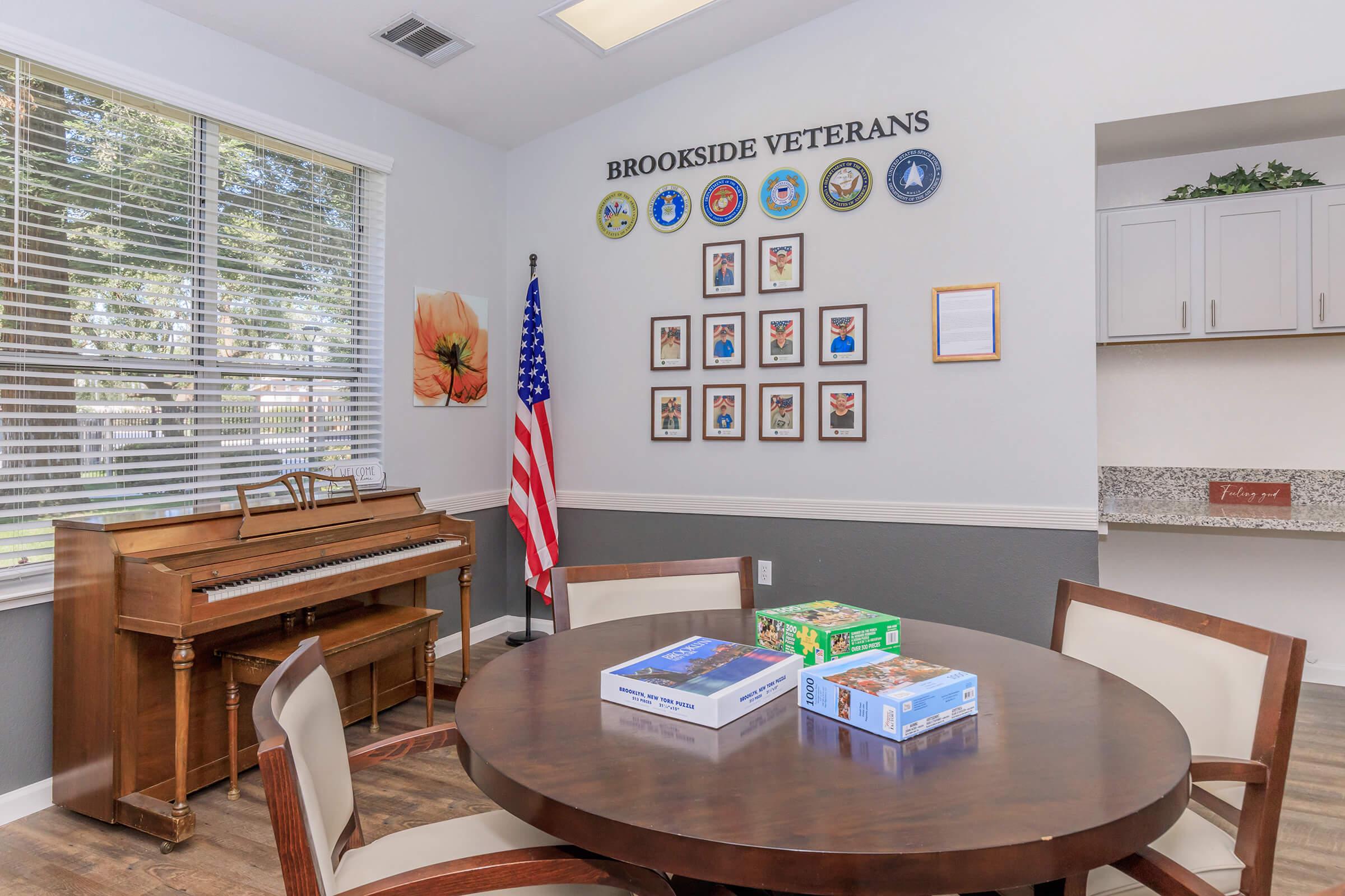 A cozy room dedicated to veterans, featuring a wooden piano and a round table with puzzle boxes. The walls display a collection of framed military honors and a flag of the United States. Large windows allow natural light to fill the space, creating a welcoming atmosphere.