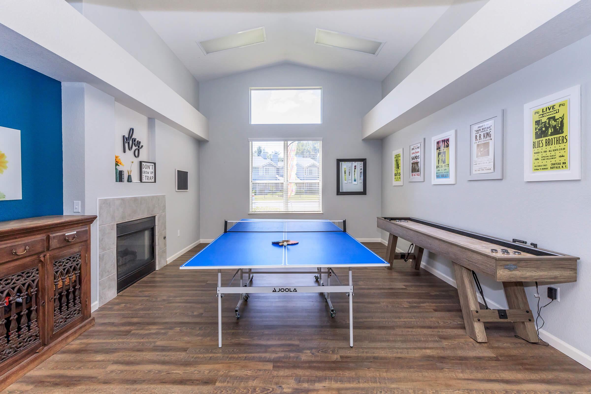 A spacious room featuring a blue ping pong table in the center, surrounded by natural light from large windows. The walls are adorned with framed posters, while a wooden side table rests against one wall. A modern fireplace adds to the cozy atmosphere, and the flooring is made of rich wood.
