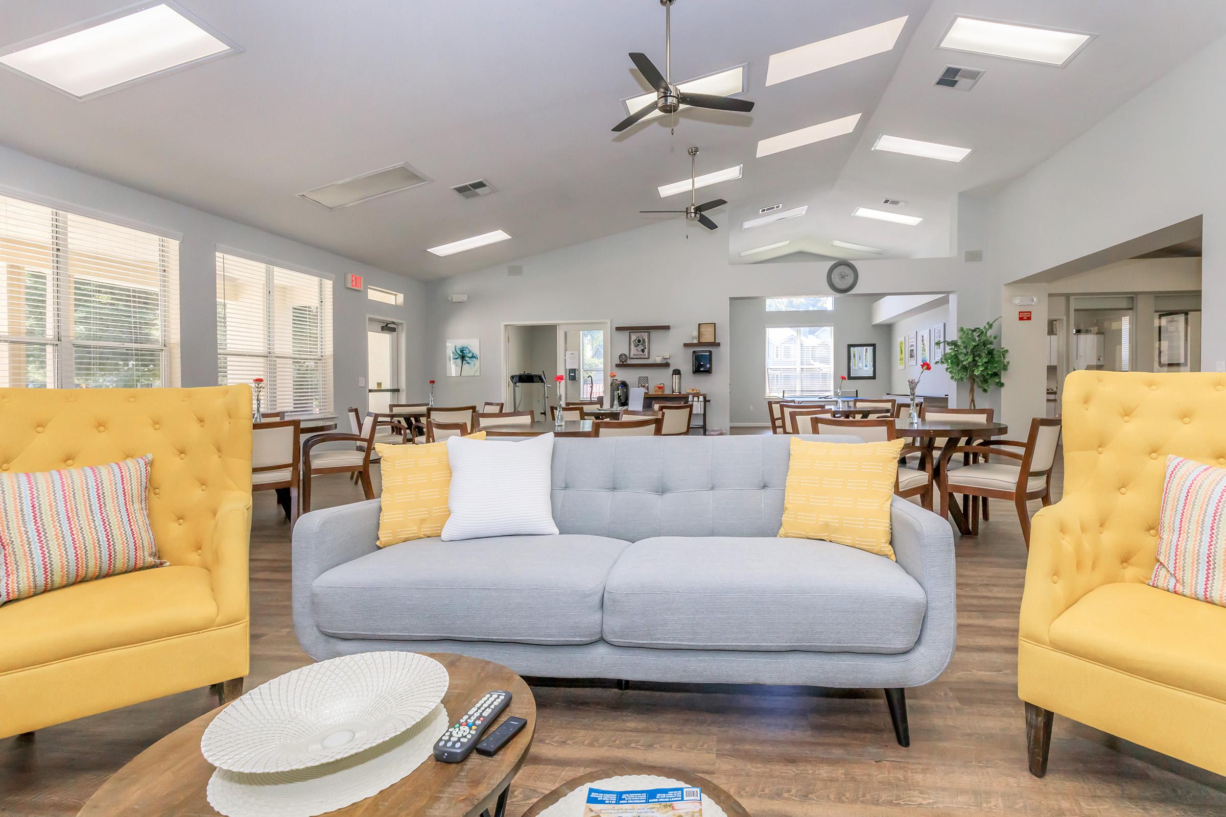 Bright and spacious common area featuring a light gray sofa with colorful accent pillows, two yellow tufted armchairs, and a wooden coffee table. Large windows allow natural light to fill the room, which has a ceiling fan and a welcoming atmosphere, with dining tables visible in the background.