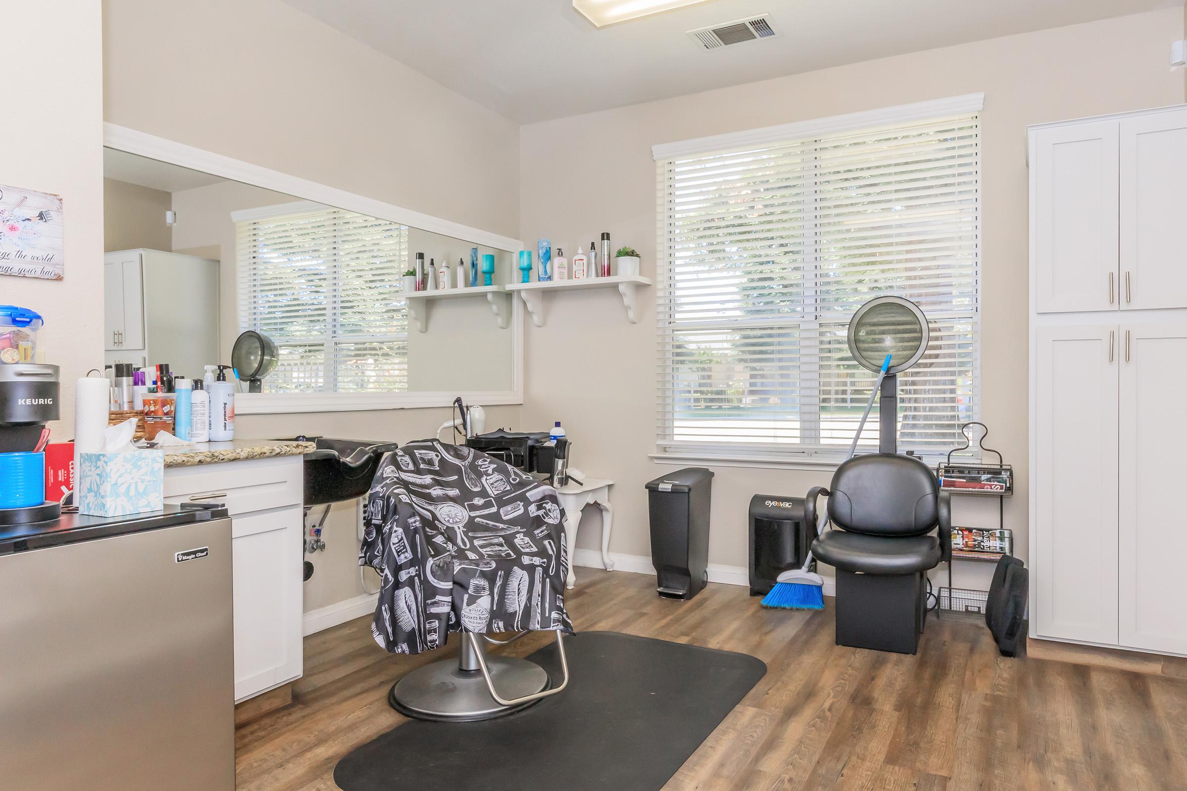 A well-lit hair salon featuring a styling chair covered with a patterned cape, a mirror along one wall, shelves with various hair products, a black rolling chair, and cleaning supplies. The space has a modern and tidy appearance with large windows allowing natural light.