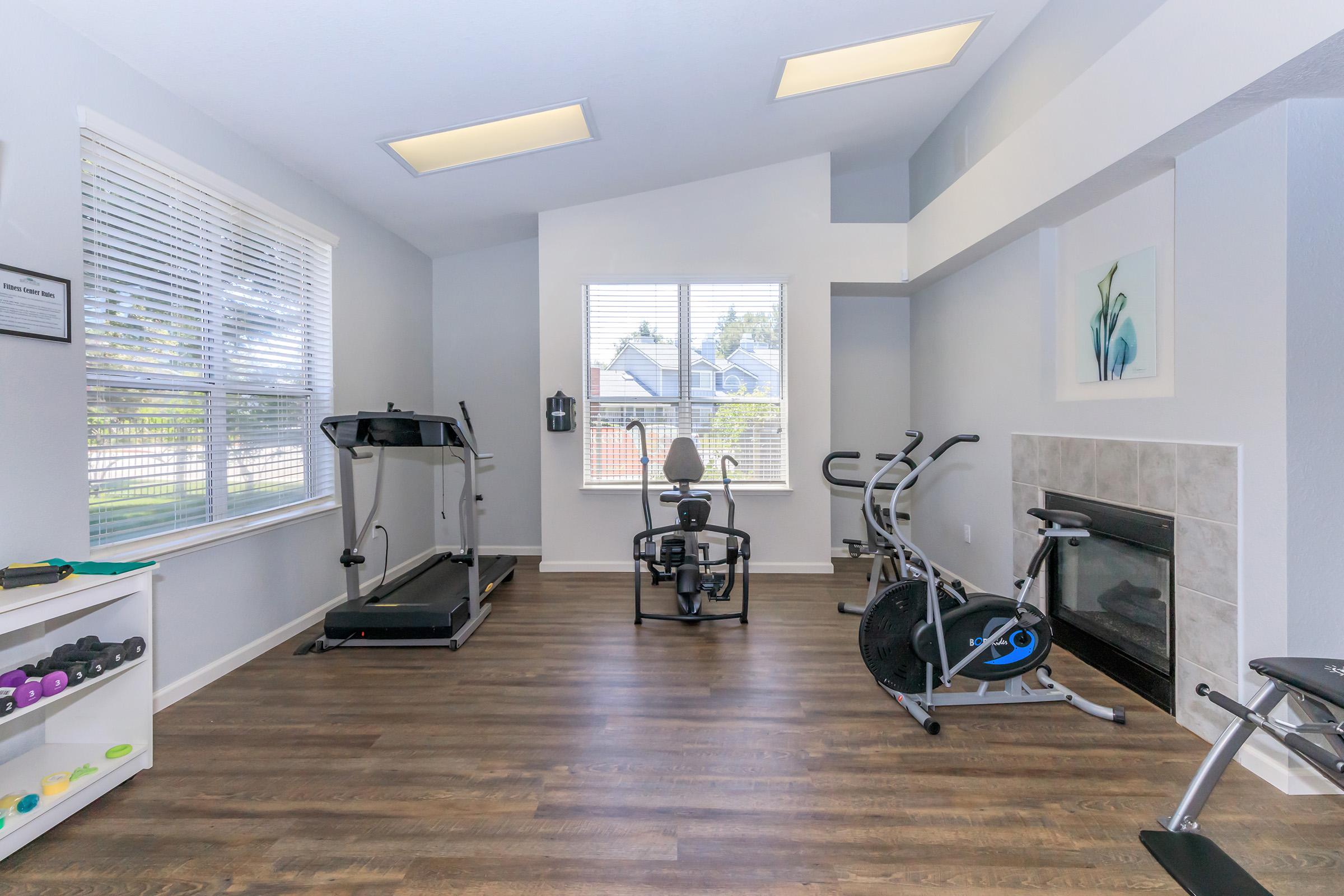 A bright home gym featuring a treadmill, exercise bike, and elliptical machine. Light-colored walls and wooden flooring enhance the airy feel. A window allows natural light, and a small fireplace adds warmth. Weights are neatly arranged on a shelf.