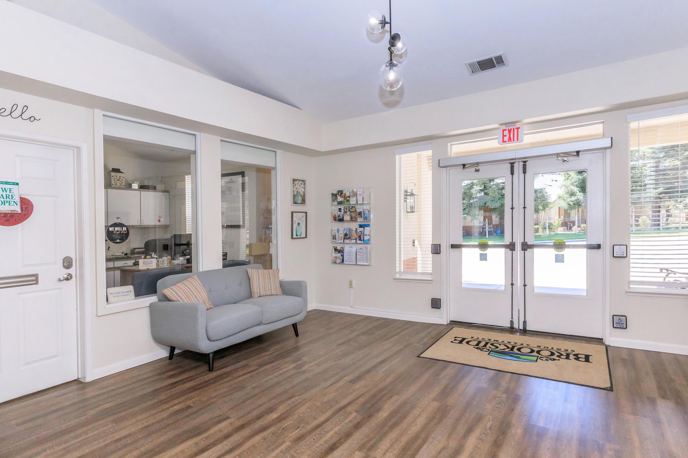 A bright and welcoming entryway featuring a light gray couch, large windows with natural light, and double glass doors leading outside. The walls are adorned with various framed photos and art. A doormat with the word "Welcome" is situated at the entrance, enhancing the inviting atmosphere.
