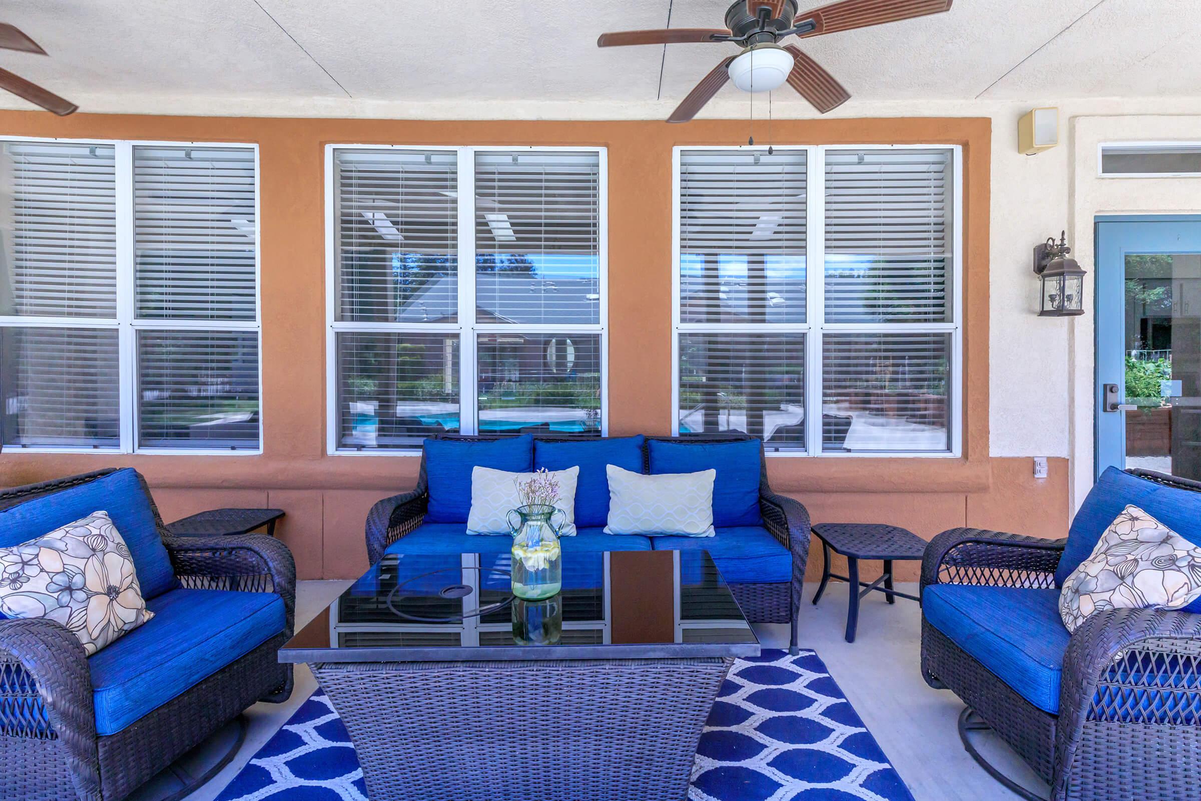 A cozy outdoor seating area featuring blue cushions on wicker furniture. There’s a glass coffee table in the center, and behind, large windows with white blinds overlook a pool area. The space is bright and welcoming, ideal for relaxation or entertaining guests.