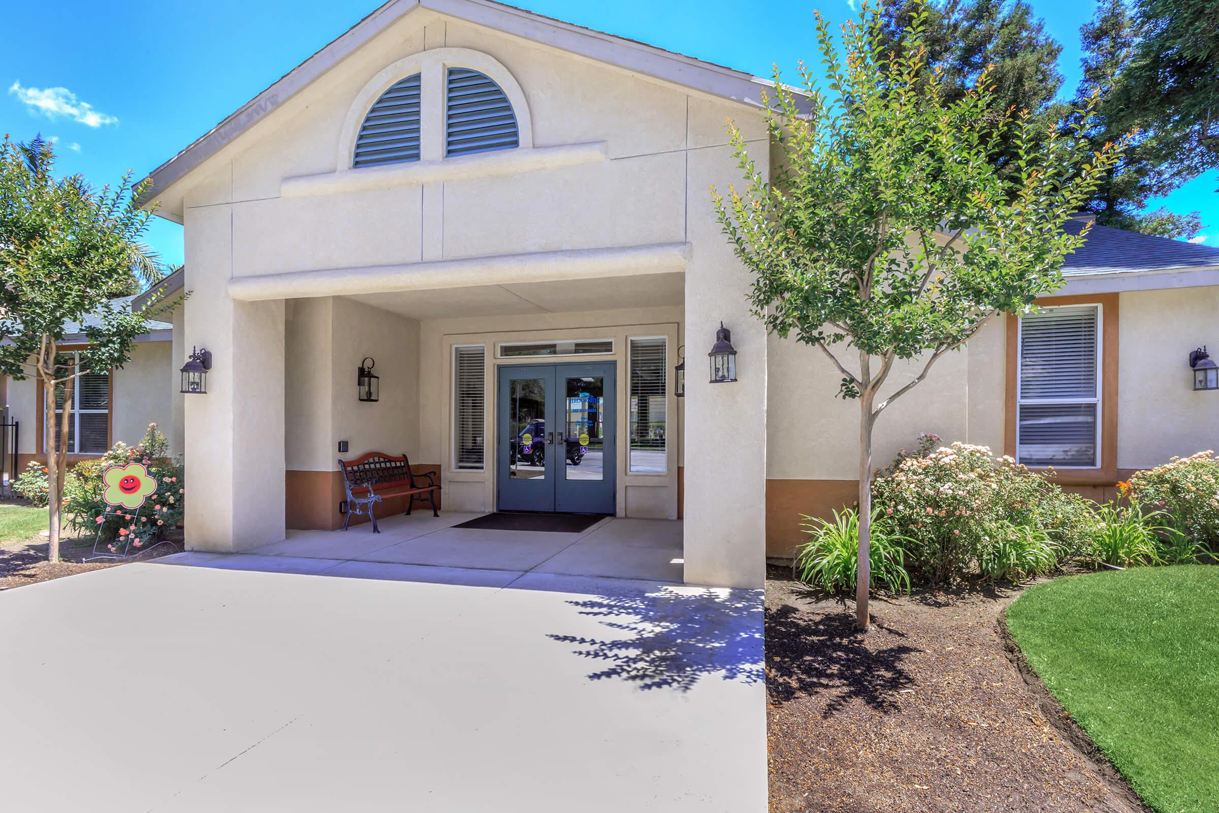 Exterior of a modern house featuring a covered porch with two front doors, flanked by decorative lanterns. The landscape includes neatly trimmed bushes and a flowering plant, with a clear blue sky in the background. A bench is positioned near the entrance, creating an inviting atmosphere.