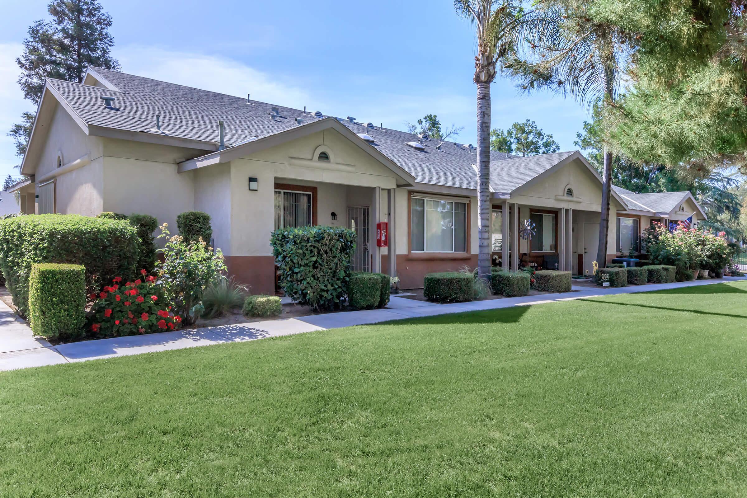 A row of single-story residential units lined with well-maintained lawns and shrubs. Each unit features a small porch, and there are vibrant flower bushes adding color to the landscape. The scene is set under a clear blue sky, with palm trees visible in the background.