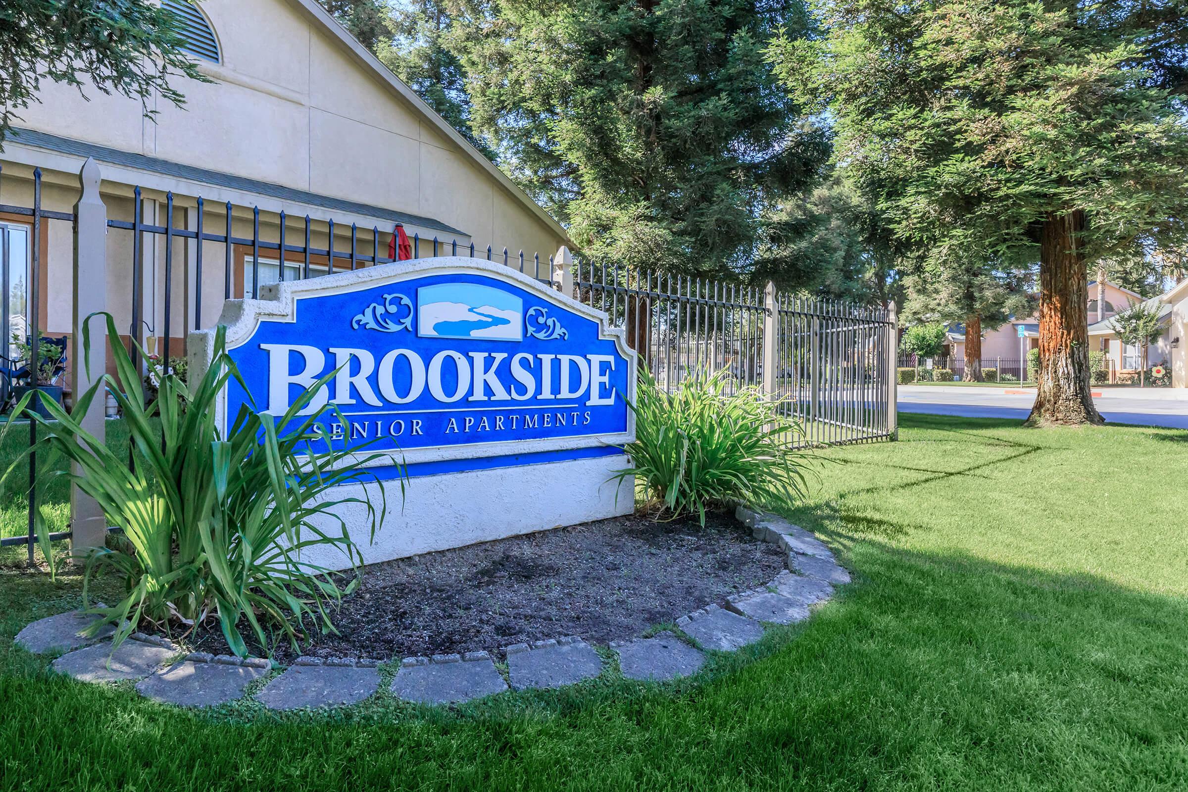 Sign for Brookside Senior Apartments surrounded by greenery and a stone pathway. The background features a residential area with trees and more buildings, indicating a peaceful community setting.
