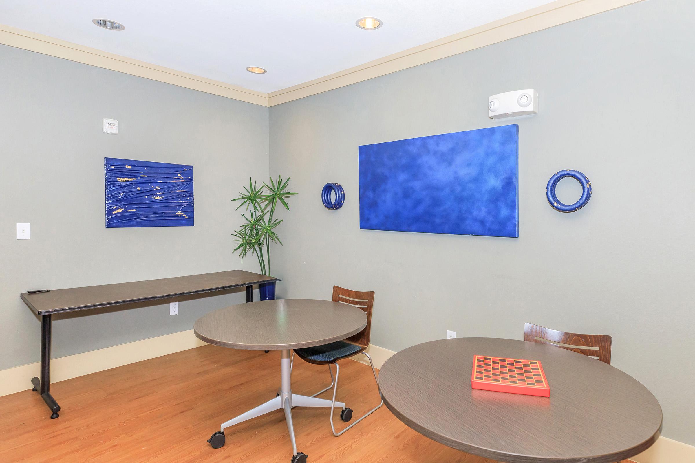 A small, modern conference room featuring two round tables, a dark wooden floor, and a green wall with abstract blue artwork. The space includes a small plant in the corner and ambient lighting, creating a calm and inviting atmosphere for meetings or discussions.