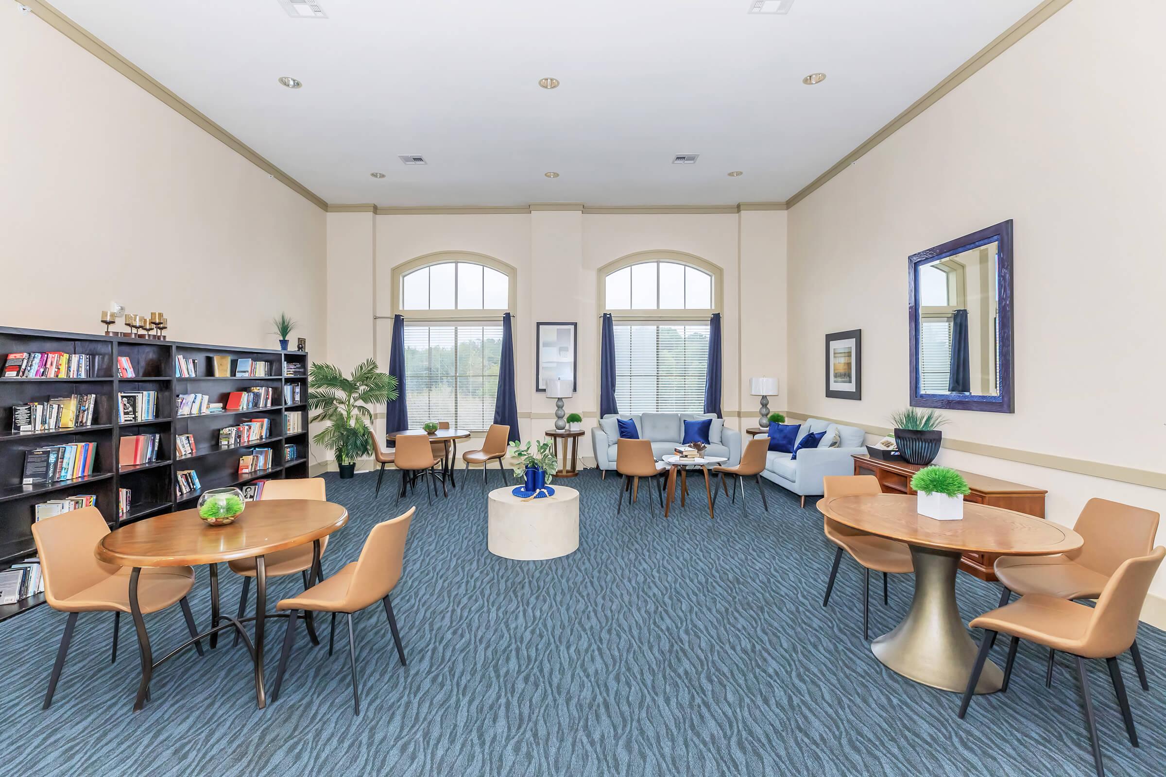 A spacious lounge area featuring several round tables with chairs, a bookshelf filled with books, and a cozy seating arrangement with blue sofas. Green plants add a touch of nature, and large windows allow natural light to brighten the room. Modern decor and a relaxed atmosphere create an inviting space for socializing or reading.