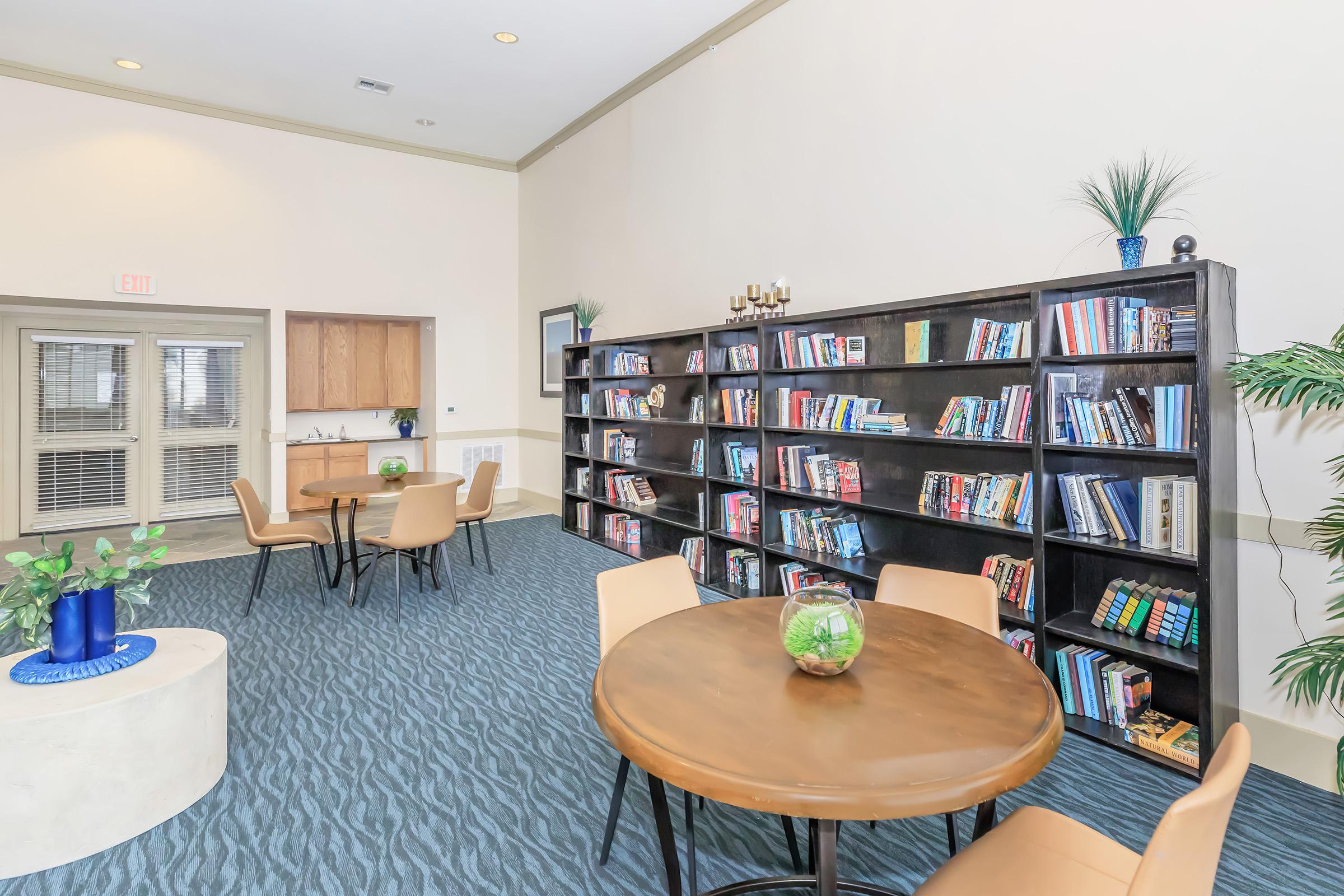A cozy reading room featuring a round wooden table with chairs, a spacious bookshelf filled with books, a small decorative plant on the table, and potted greenery. Natural light filters through the windows, creating an inviting atmosphere for relaxation and reading.