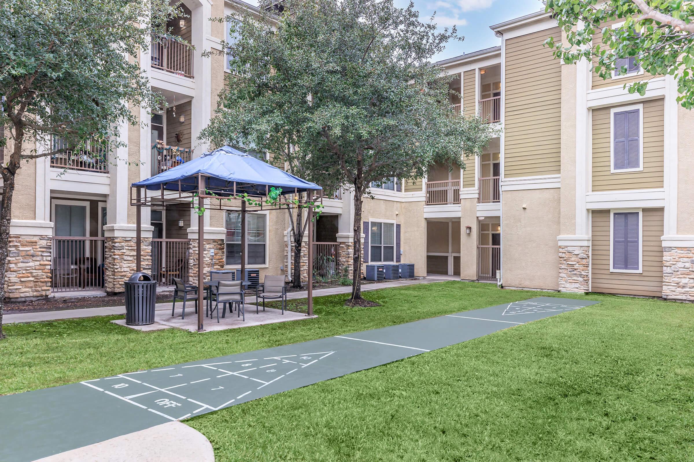 A grassy area between apartment buildings featuring a blue pavilion with seating, a path for games, and shaded trees. The scene is well-lit, showcasing a welcoming outdoor space for relaxation and activities.