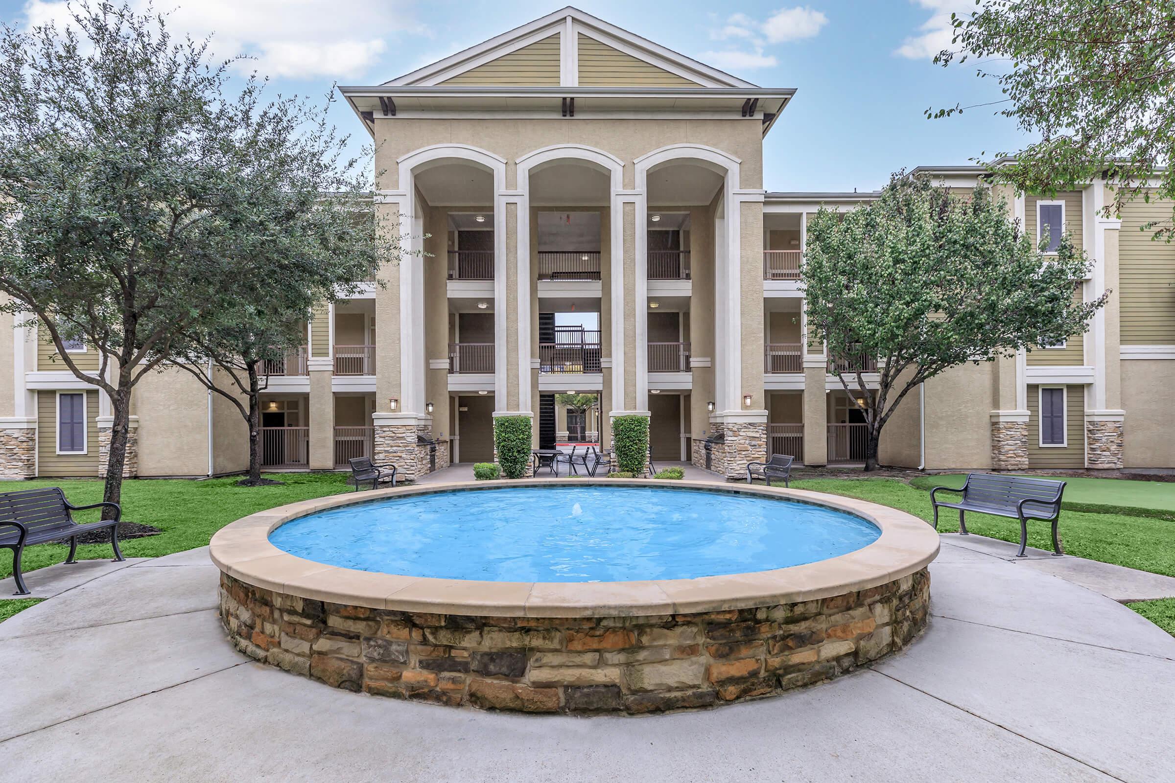 A landscaped courtyard features a circular stone fountain with blue water at the center, surrounded by benches and greenery. In the background, a multi-story building with a light-colored façade and arched entrances stands, flanked by trees. The scene conveys a tranquil, inviting atmosphere.