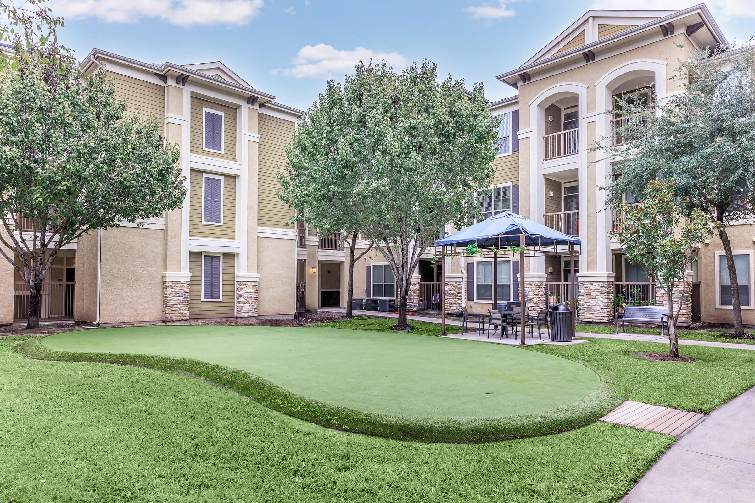 A well-maintained courtyard featuring manicured green grass, surrounded by several beige residential buildings. In the center, there’s a gazebo with seating and tables, and trees providing shade, creating a serene outdoor space.