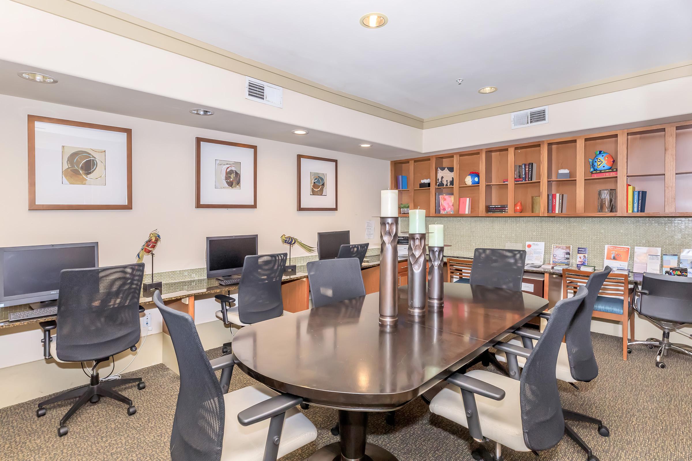 A modern office space featuring a large wooden conference table surrounded by ergonomic chairs. In the background, there are two desks equipped with computers, a bookshelf filled with books, and decorative items. The walls are adorned with framed artwork, and the overall atmosphere is professional and inviting.
