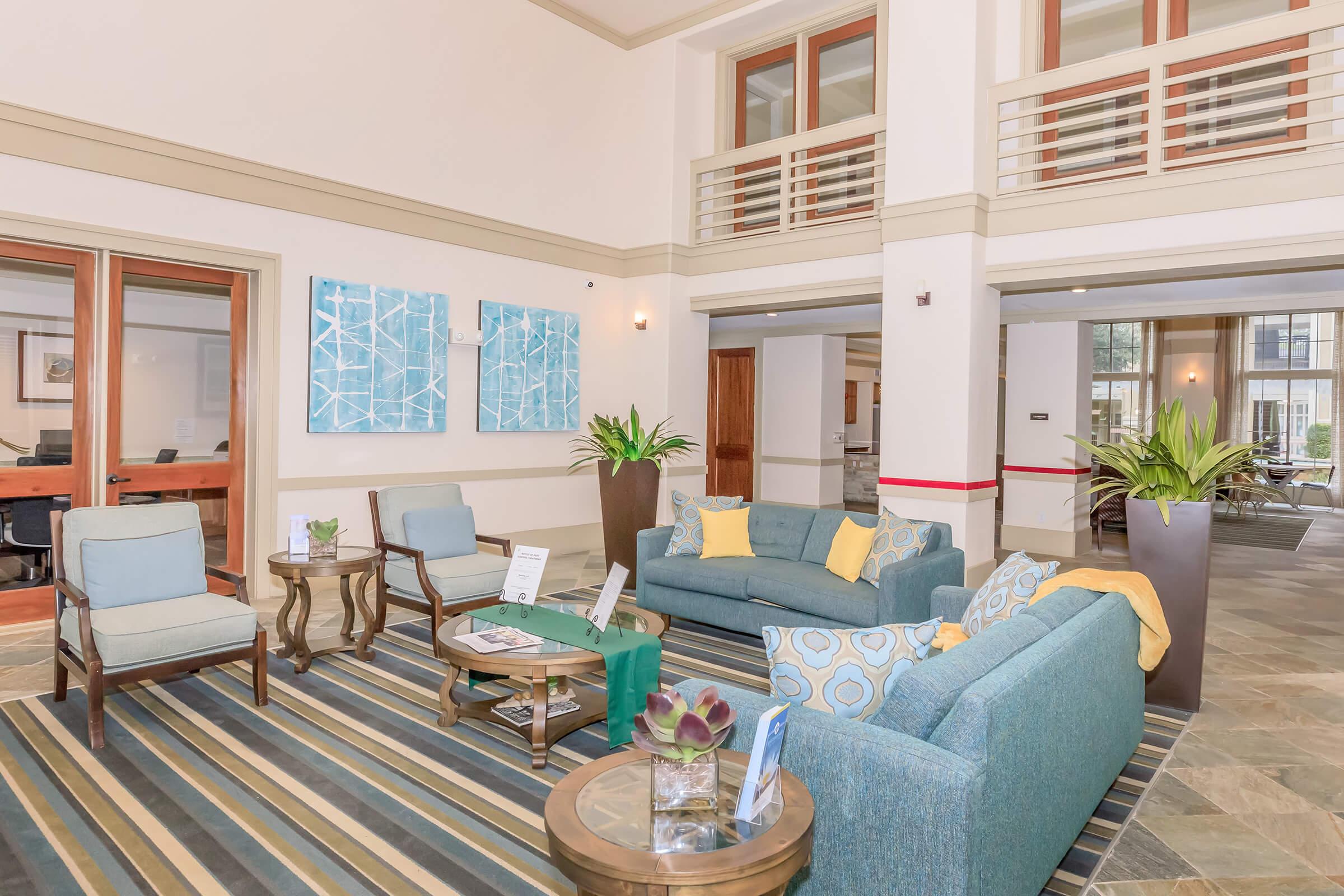 A modern lobby area featuring blue upholstered seating, including a couch and chairs, arranged around wooden tables. Decorative plants are positioned around the space, with artwork on the walls. The floor is covered with striped rugs, and large windows allow natural light to brighten the room.
