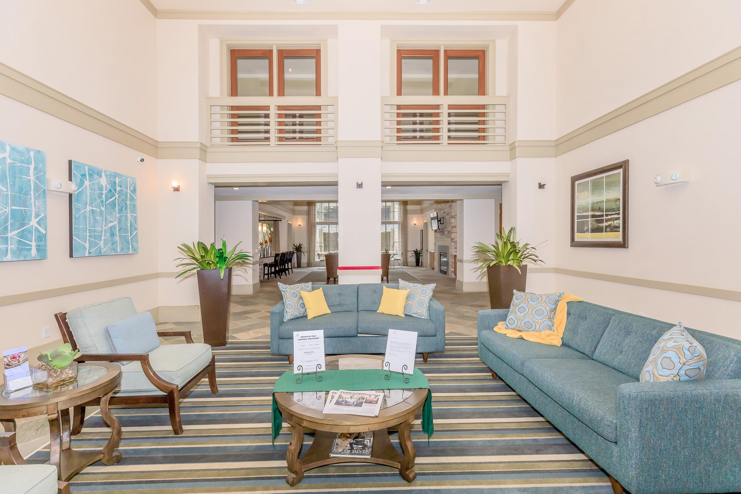 A well-lit lobby area featuring a cozy seating arrangement with a blue sofa and two armchairs. Decorative cushions add color, while a coffee table holds informational brochures. Lush green plants and artworks adorn the walls, and large windows provide natural light in a welcoming atmosphere.