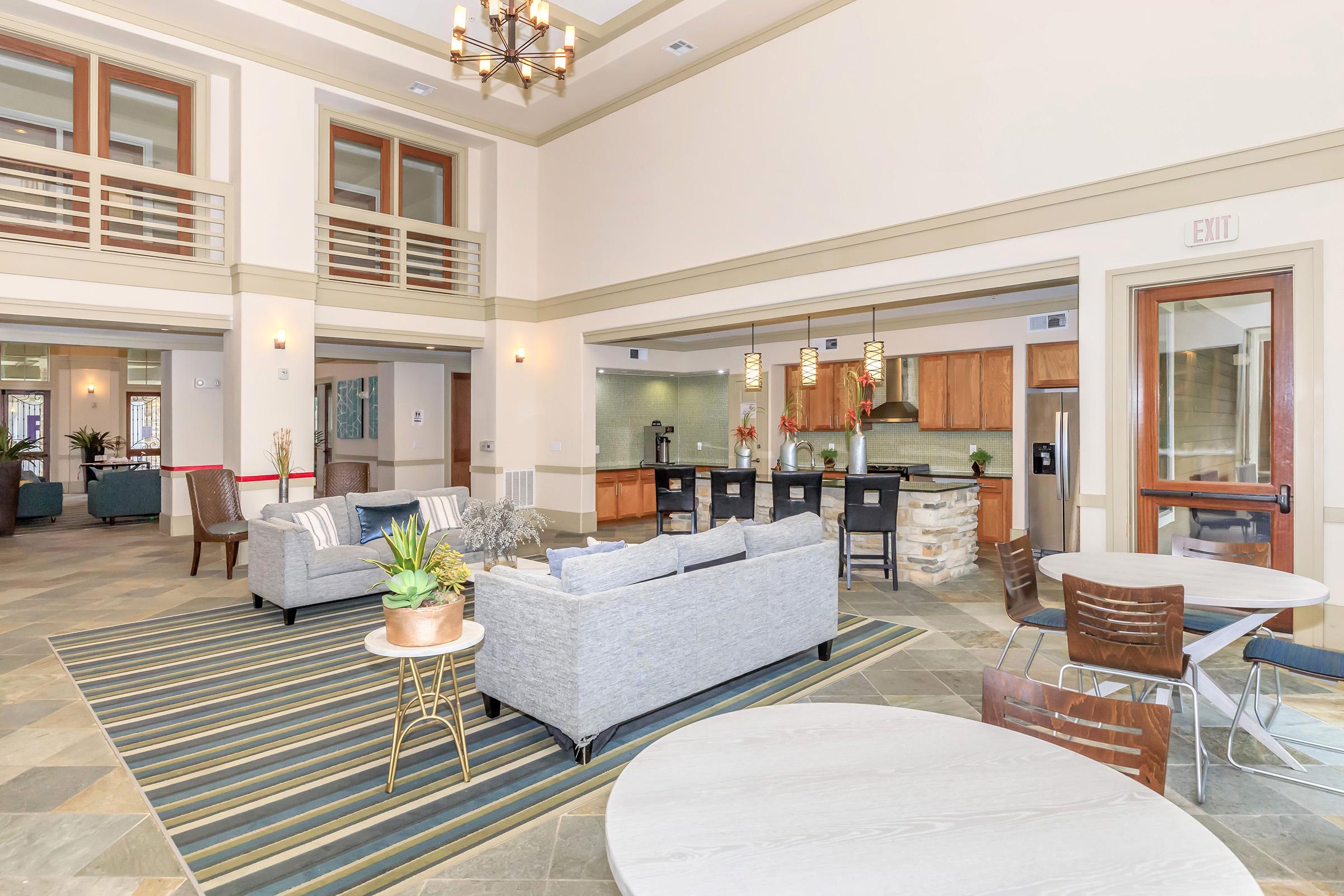 Interior view of a spacious, modern common area featuring a seating arrangement with gray sofas, a coffee table, and decorative plants. The room has large windows with natural light, warm-toned cabinetry in the kitchen area, and a contemporary dining setup with round tables and wooden chairs on a patterned rug.