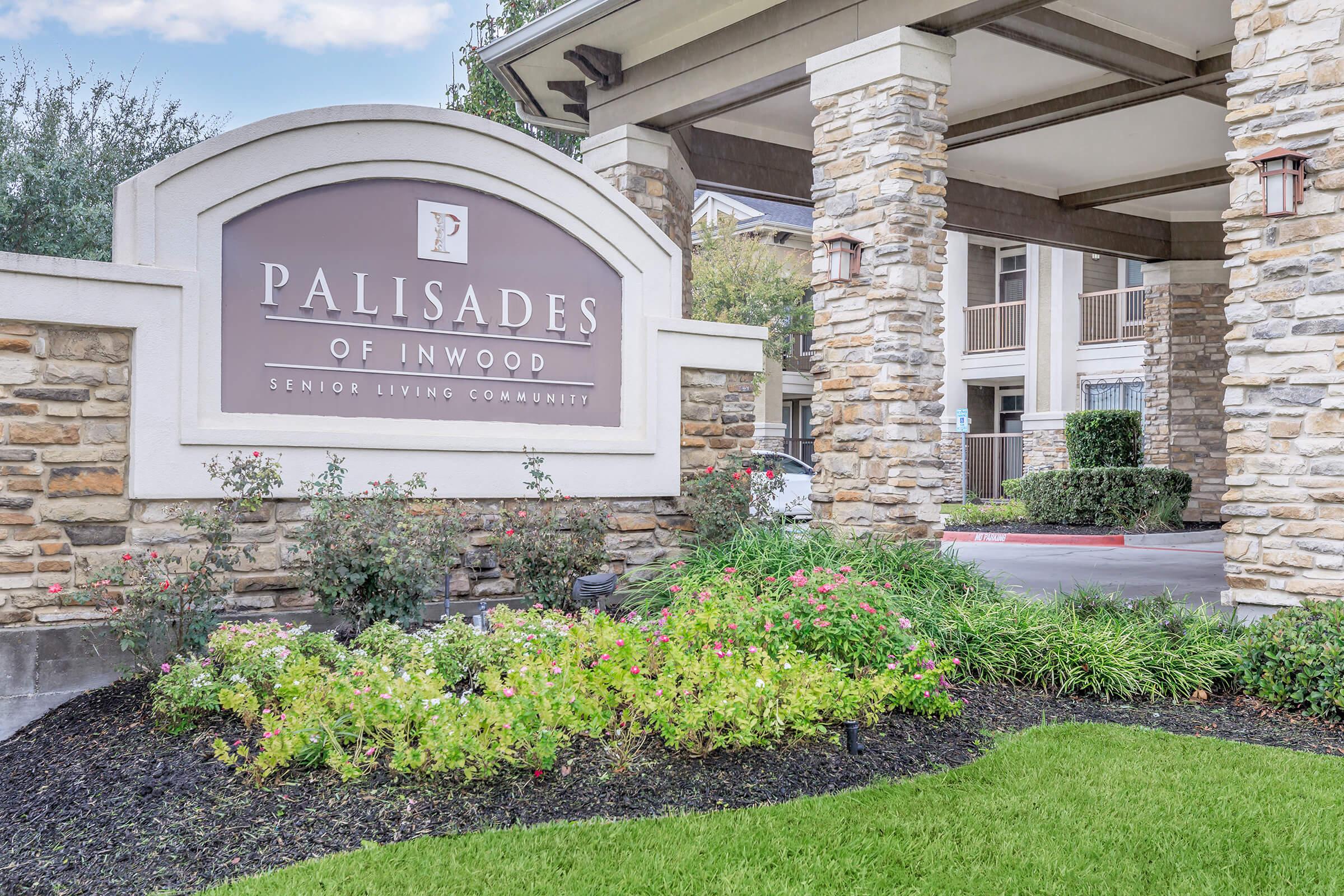Sign for "Palisades of Inwood" Senior Living Community, featuring a stone facade with a landscaped area of flowers and greenery in front. The building has a welcoming appearance, with a modern entrance and surrounding trees.