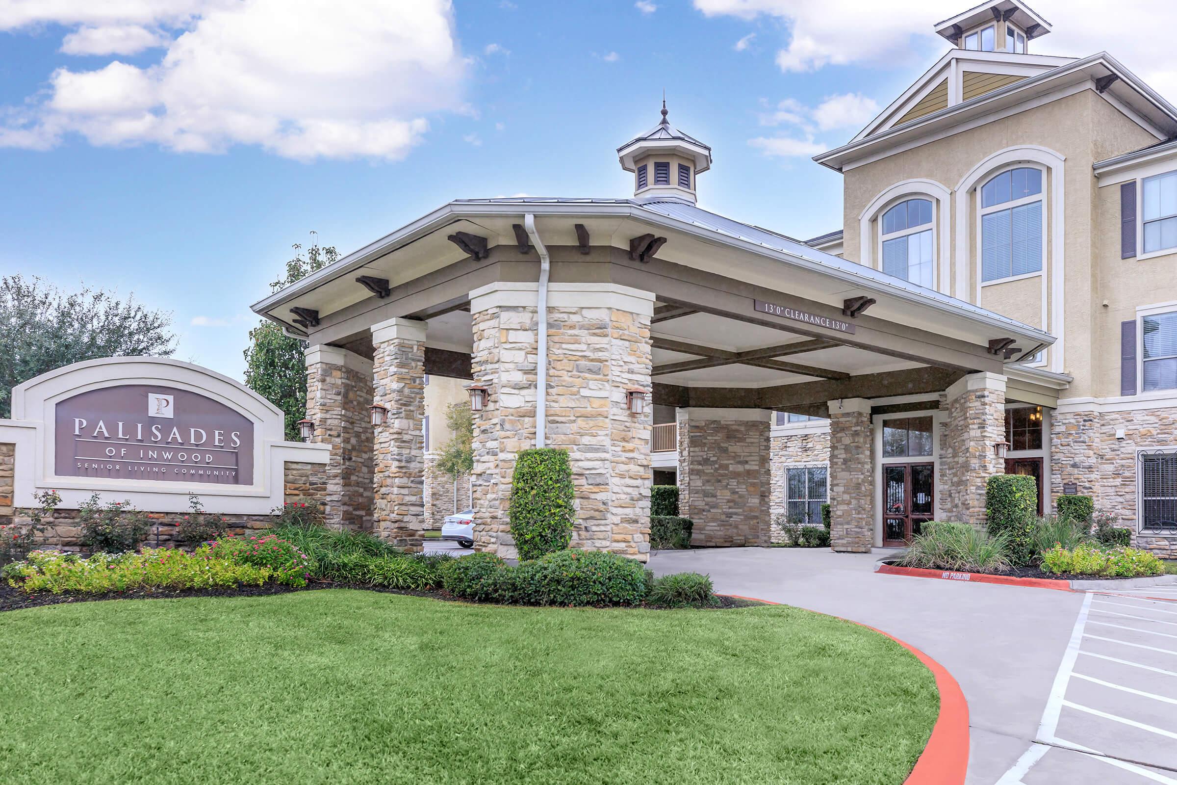 Entrance to the Palisades of Inwood, a residential building, featuring a stone-covered portico supported by pillars. The signage is prominently displayed. Lush green landscaping surrounds the entrance, with a clear blue sky in the background, creating a welcoming atmosphere.
