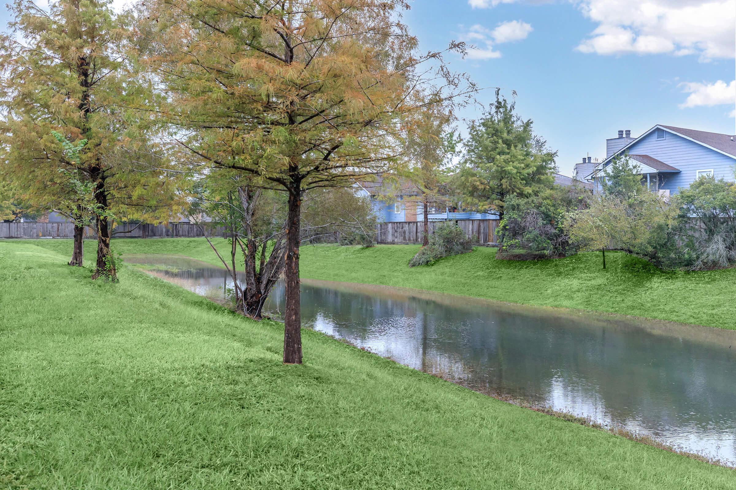 A serene landscape featuring a calm, reflective river bordered by lush green grass and trees with autumn foliage. In the background, residential houses can be seen, framed by clear blue skies and fluffy clouds, creating a peaceful suburban atmosphere.