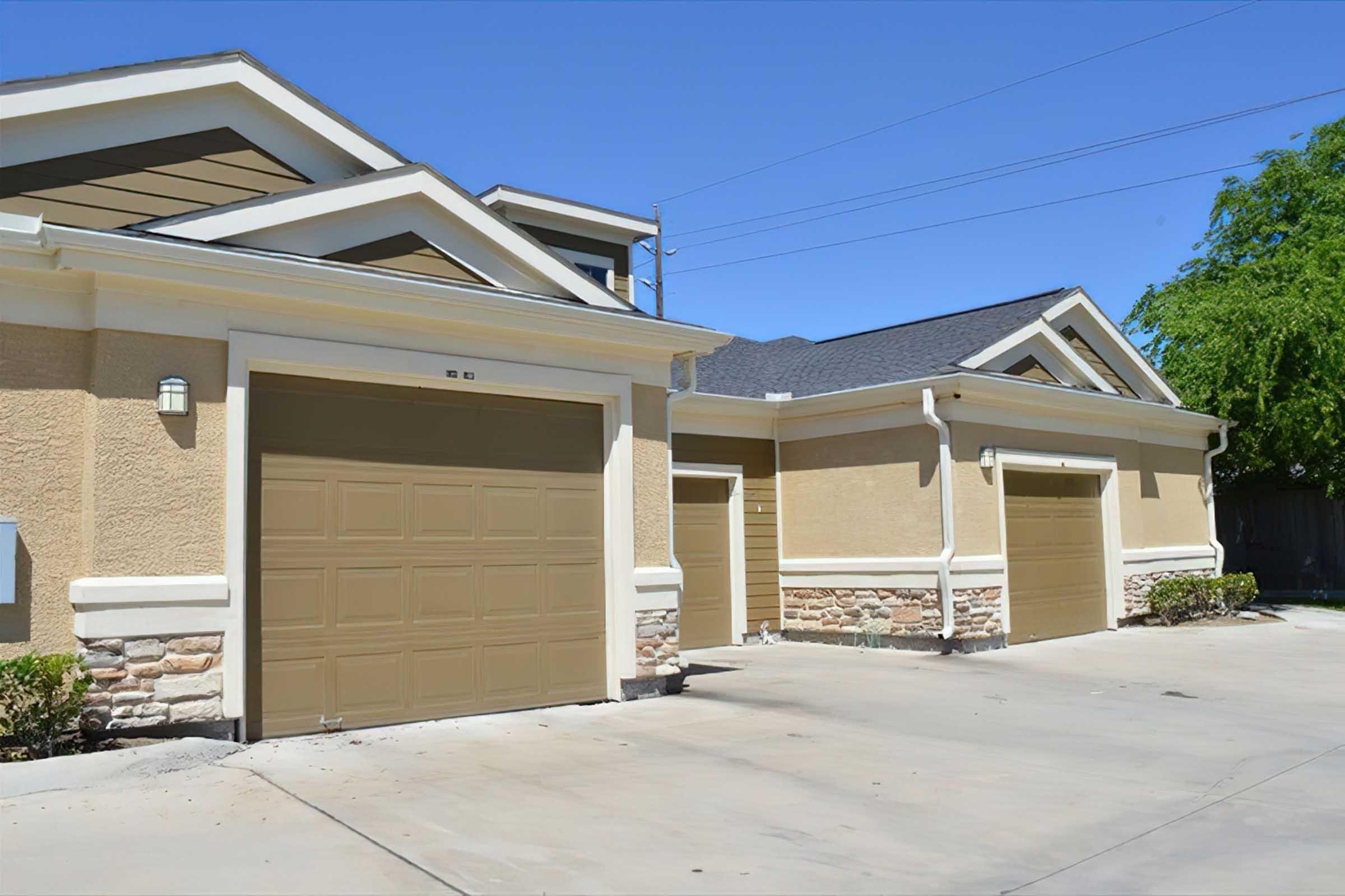 A view of a residential garage with two beige garage doors, set against a light-colored exterior wall. The garage is part of a larger house featuring a sloped roof and landscaped area, with clear blue skies overhead.