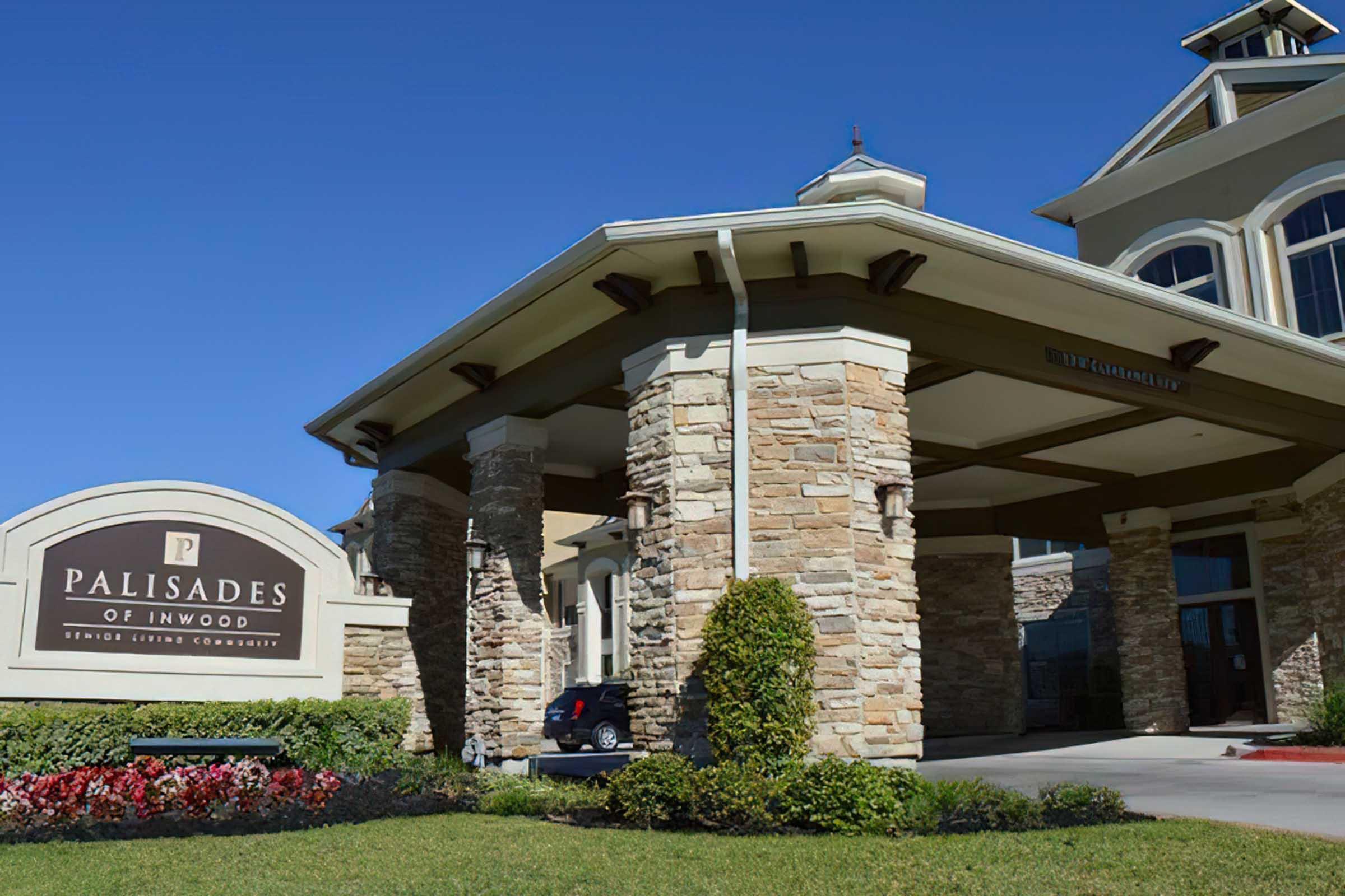 Entrance of a senior living community named "Palisades of Inwood," featuring stone pillars and a covered entryway. The sign is prominently displayed, and there is blue sky in the background. Green grass surrounds the building, and a car is parked nearby.