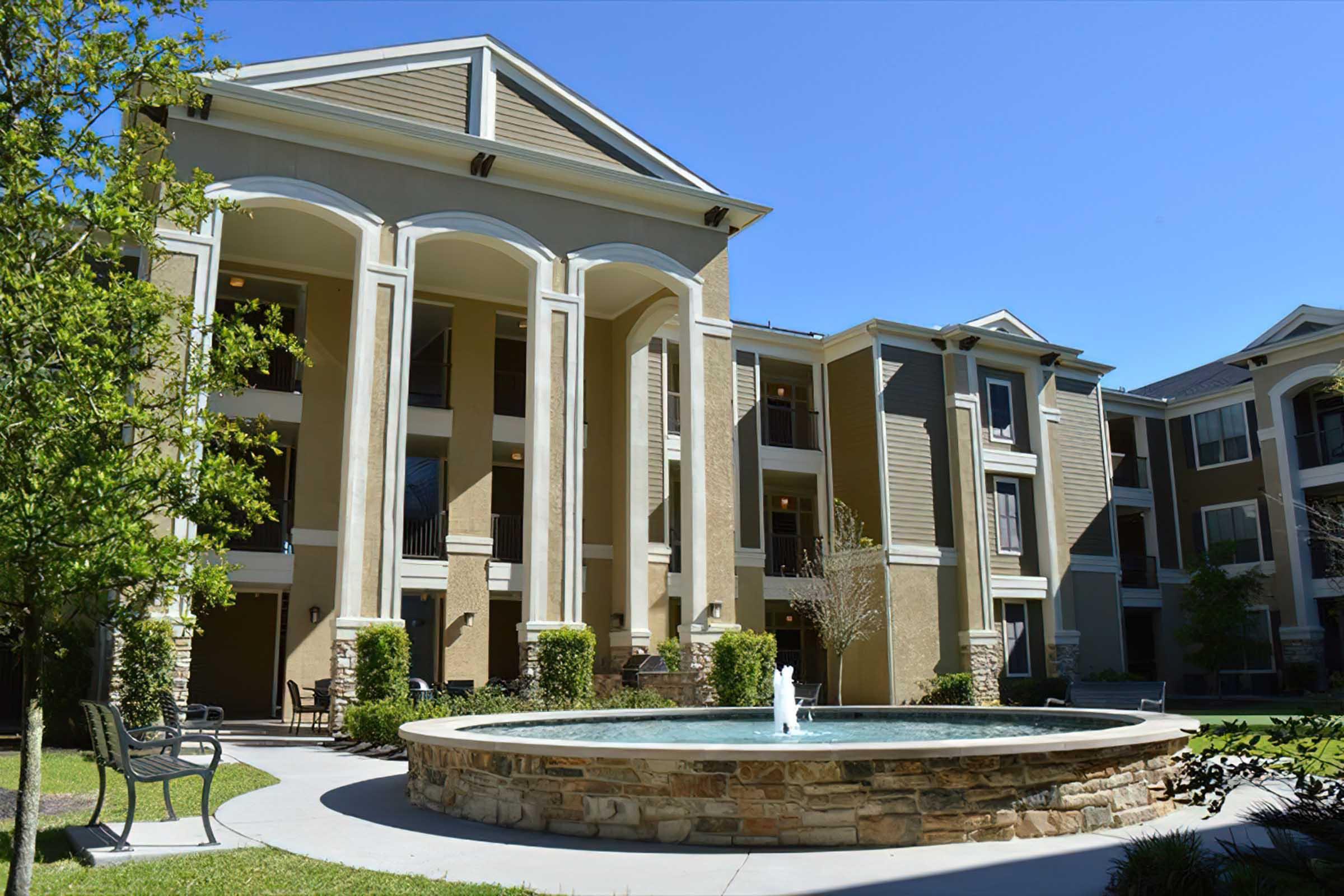 A modern apartment complex features a central fountain surrounded by greenery and seating. The building has multiple stories with balconies and large windows, set against a clear blue sky.